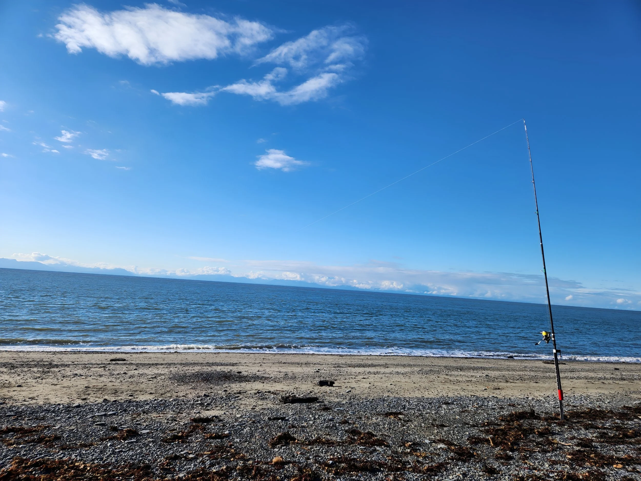 A fishing rod planted in the sandy beach facing the ocean under a partly cloudy blue sky.