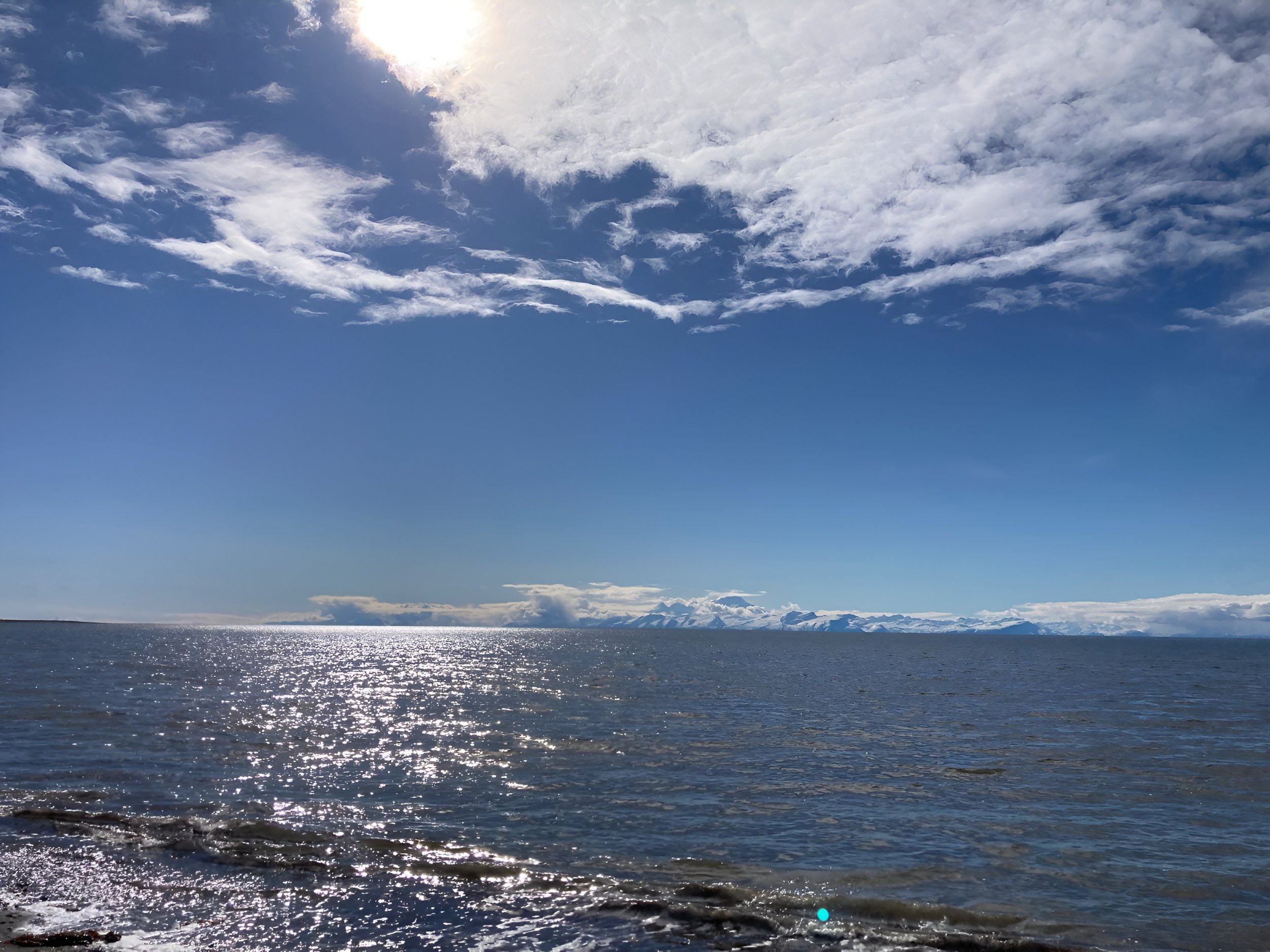 A large body of water, possibly a lake or ocean, with the sun reflecting off the surface. The sky is mostly cloudy with some patches of blue and distant snow-capped mountains on the horizon.