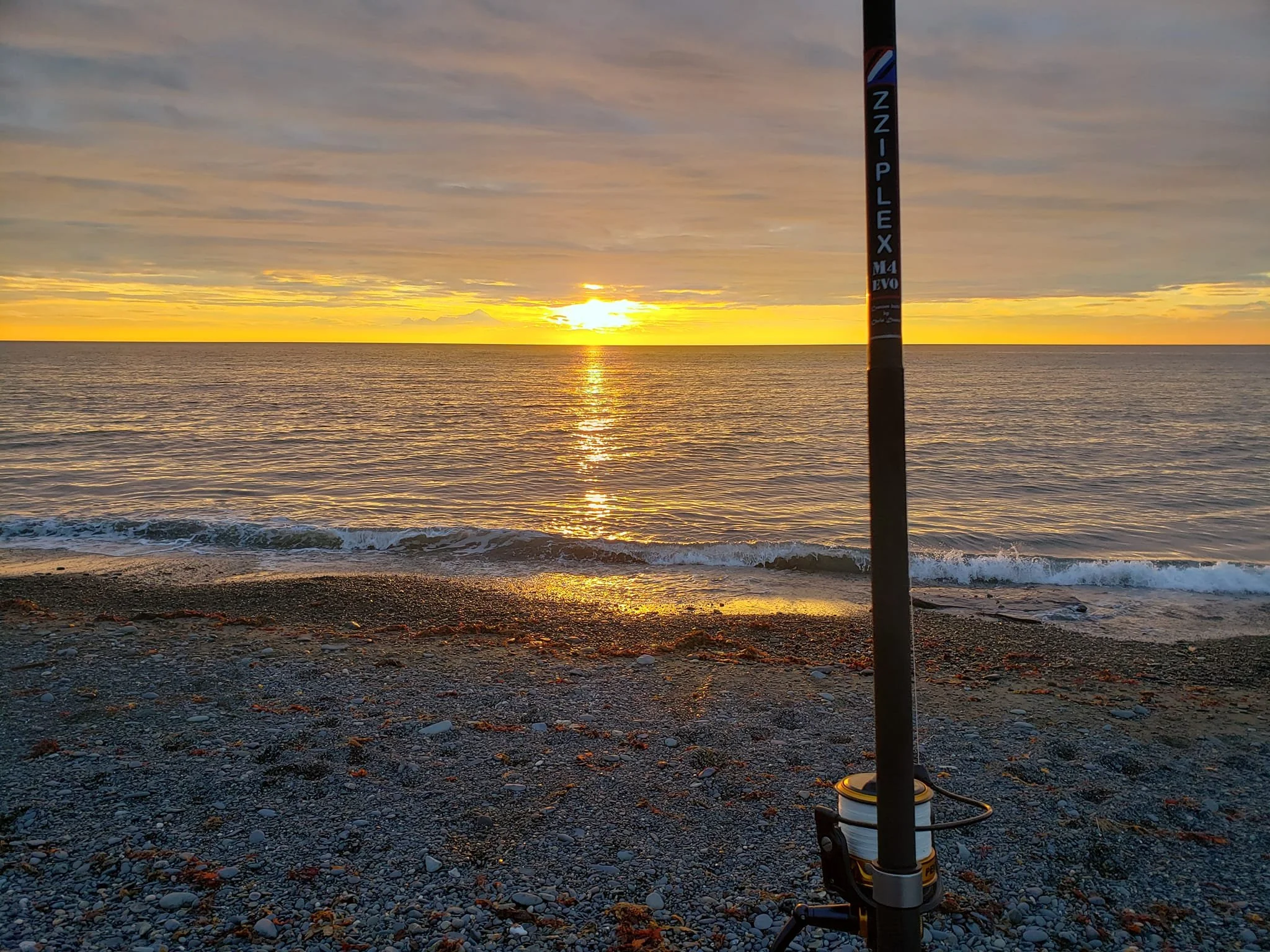 A fishing pole standing on a pebbled beach during a sunset over the ocean.