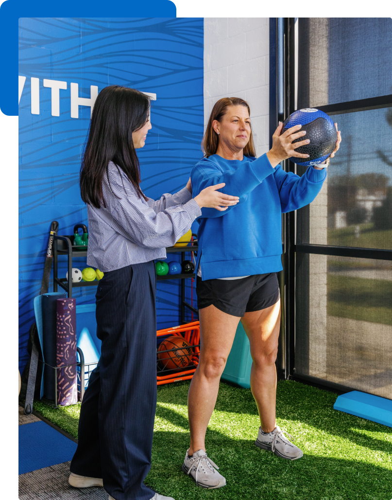 A woman in a blue sweatshirt and black shorts is holding a weighted medicine ball, while a woman in a striped shirt and business pants assists her in a fitness training session near a window at a gym.