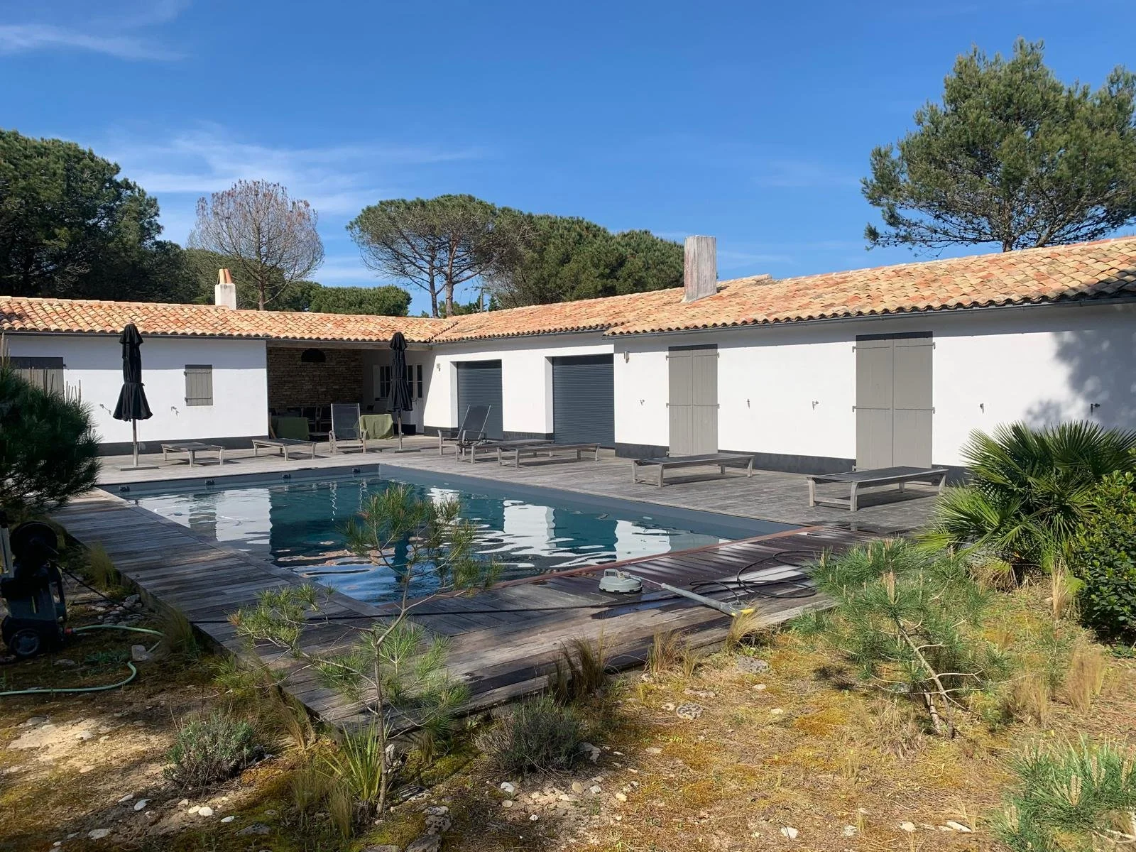 Cour intérieure avec piscine, chaises longues, parasols, bâtiment blanc avec portes grises, arbres verts et ciel bleu.