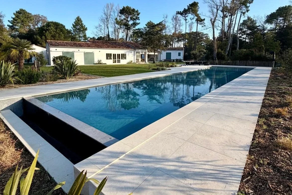 Piscine rectangulaire dans un jardin bordée de dalles blanches, avec une maison blanche et des arbres en arrière-plan.