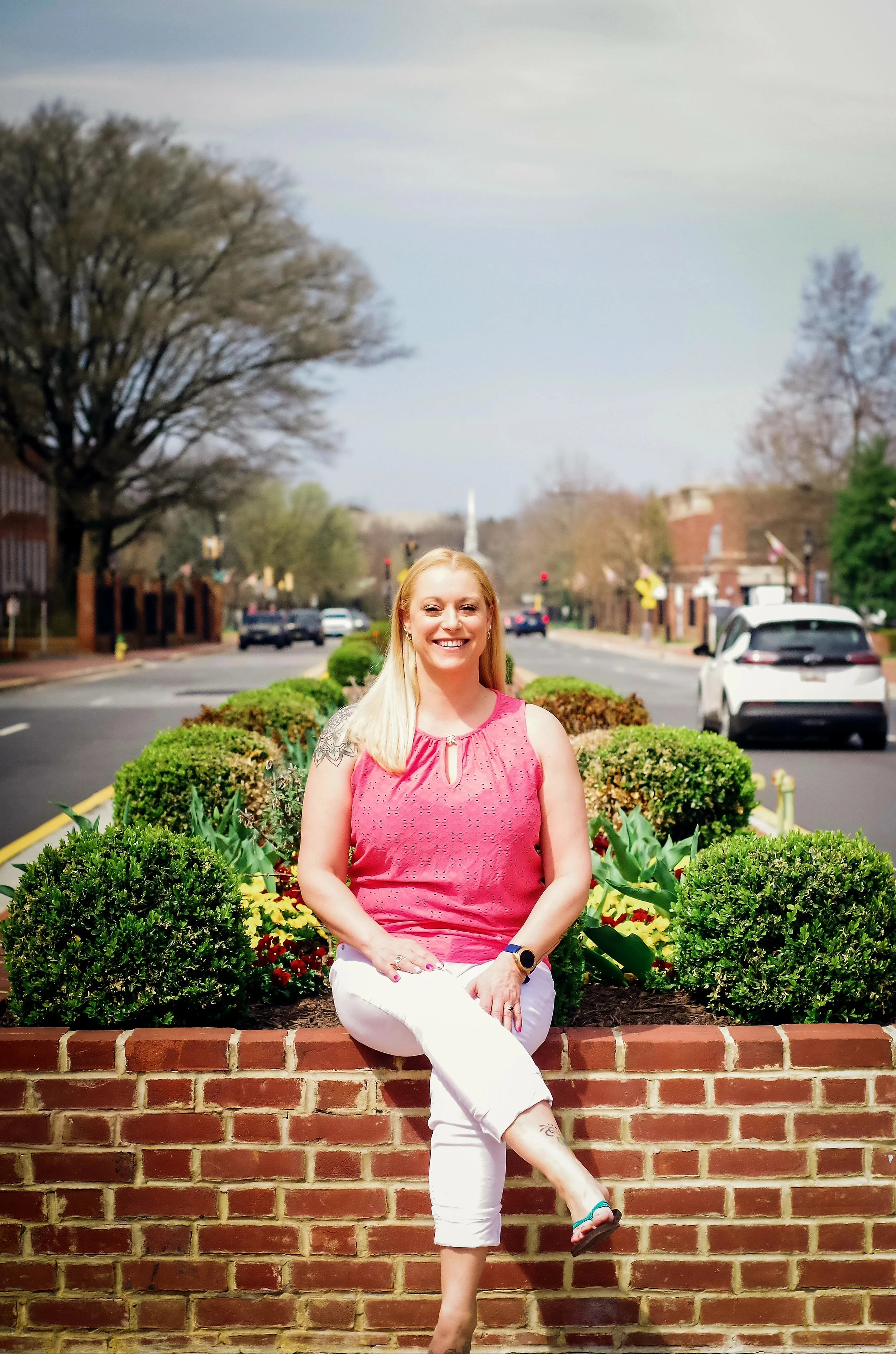 A woman sitting on a brick ledge in front of a flower bed on a city street, smiling at the camera.