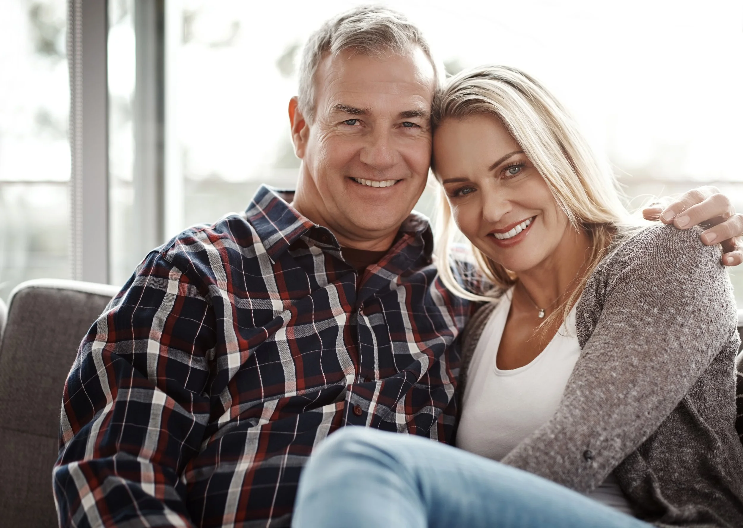 A smiling middle-aged man and woman sitting closely together on a couch, with the man wearing a plaid shirt and the woman wearing a white top and gray cardigan, in a well-lit room with large windows in the background.