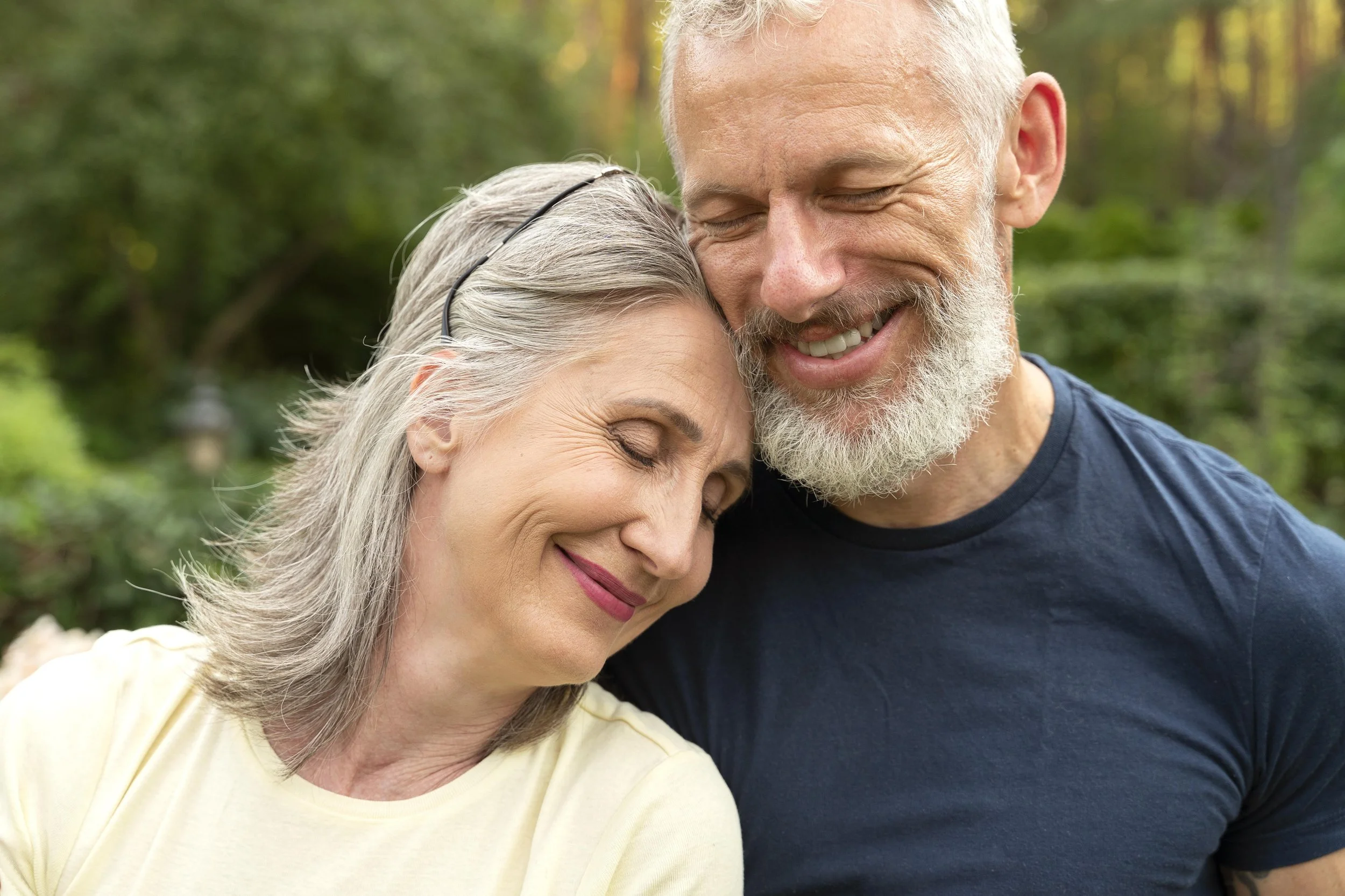 An elderly couple enjoying a tender moment outdoors, with their heads touching and smiling.