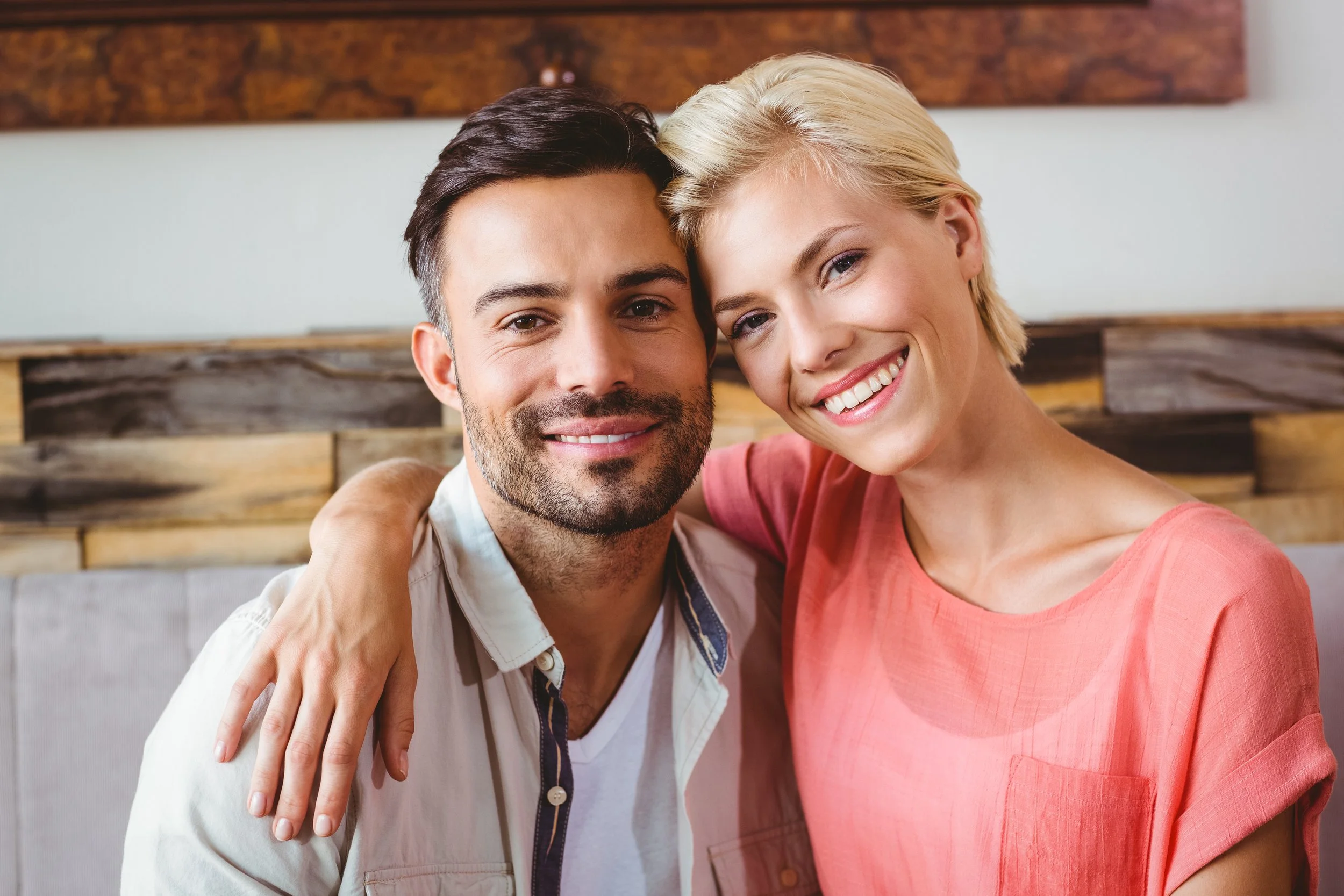 A happy couple sitting close together, smiling, with the woman's arm around the man's shoulder in a cozy indoor setting.