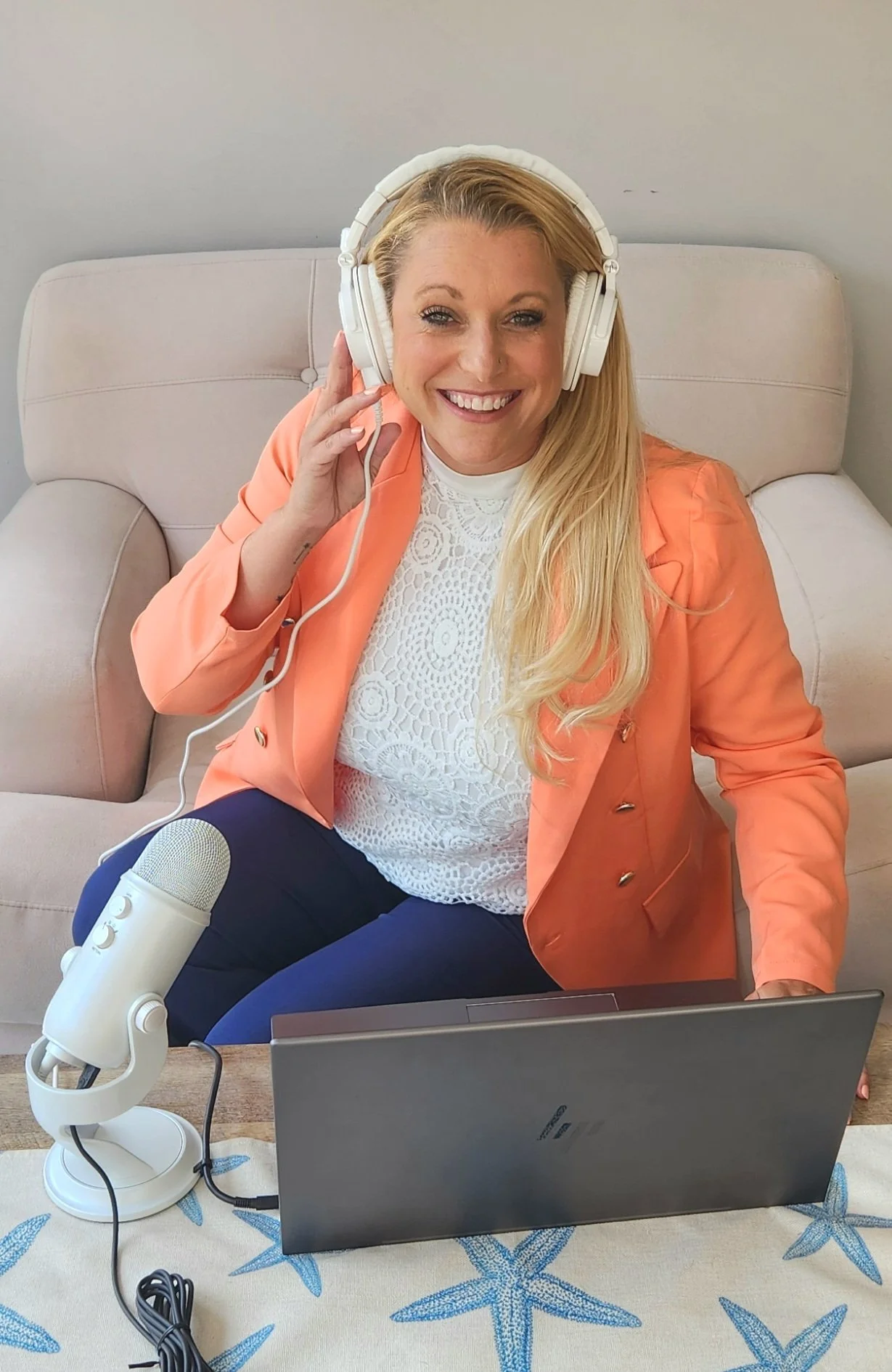 A woman with long blonde hair sitting on a beige couch, smiling, wearing a peach-colored blazer and a white lace top, holding a pair of large white headphones to her ears, with a microphone and laptop on the table in front of her.