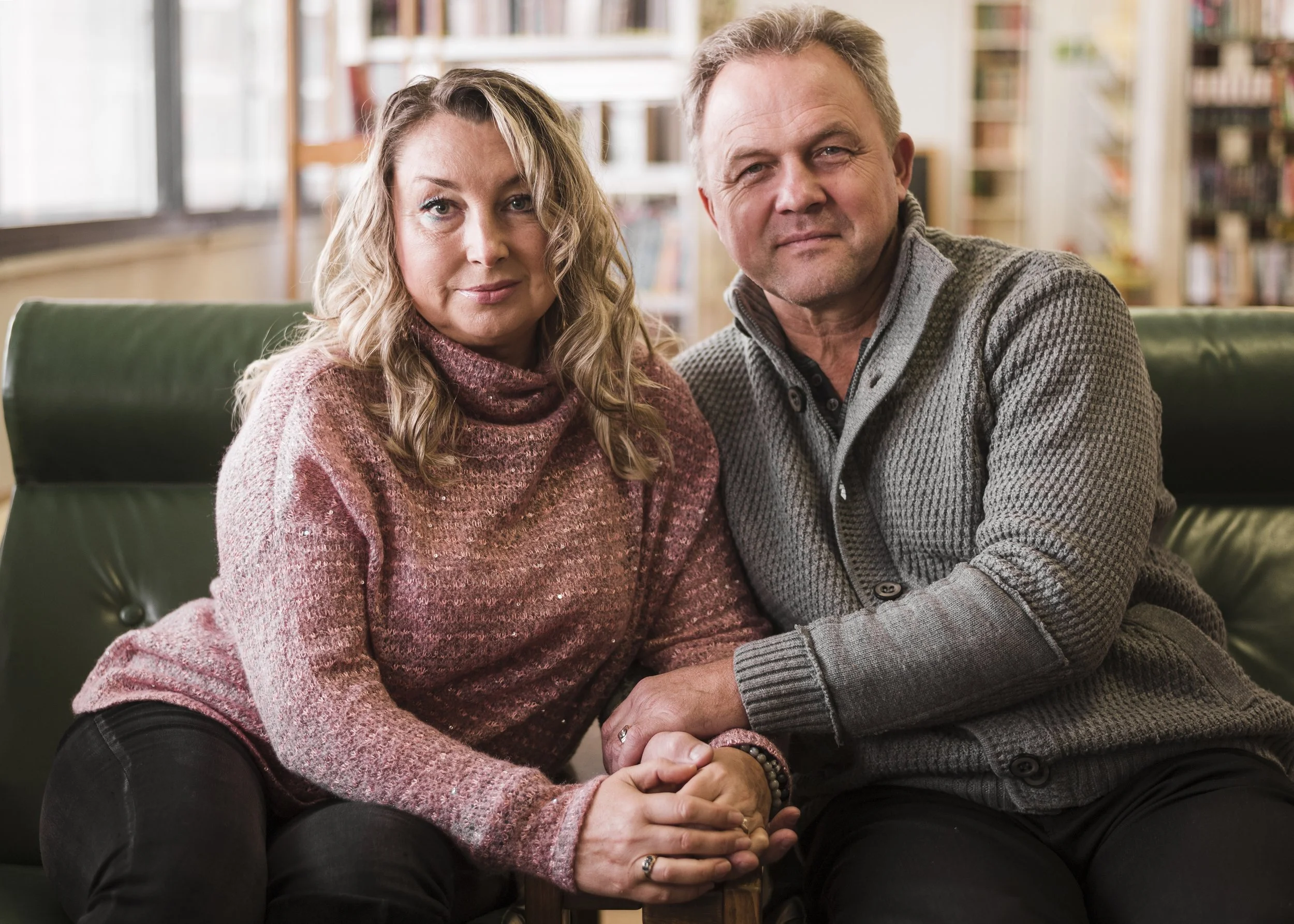 A middle-aged couple sitting close together on a green couch in a cozy room, holding hands and looking at the camera. The woman has wavy blonde hair and wears a pink sweater, and the man has short light brown hair and wears a gray sweater. The background features a well-stocked bookshelf and large windows letting in natural light.