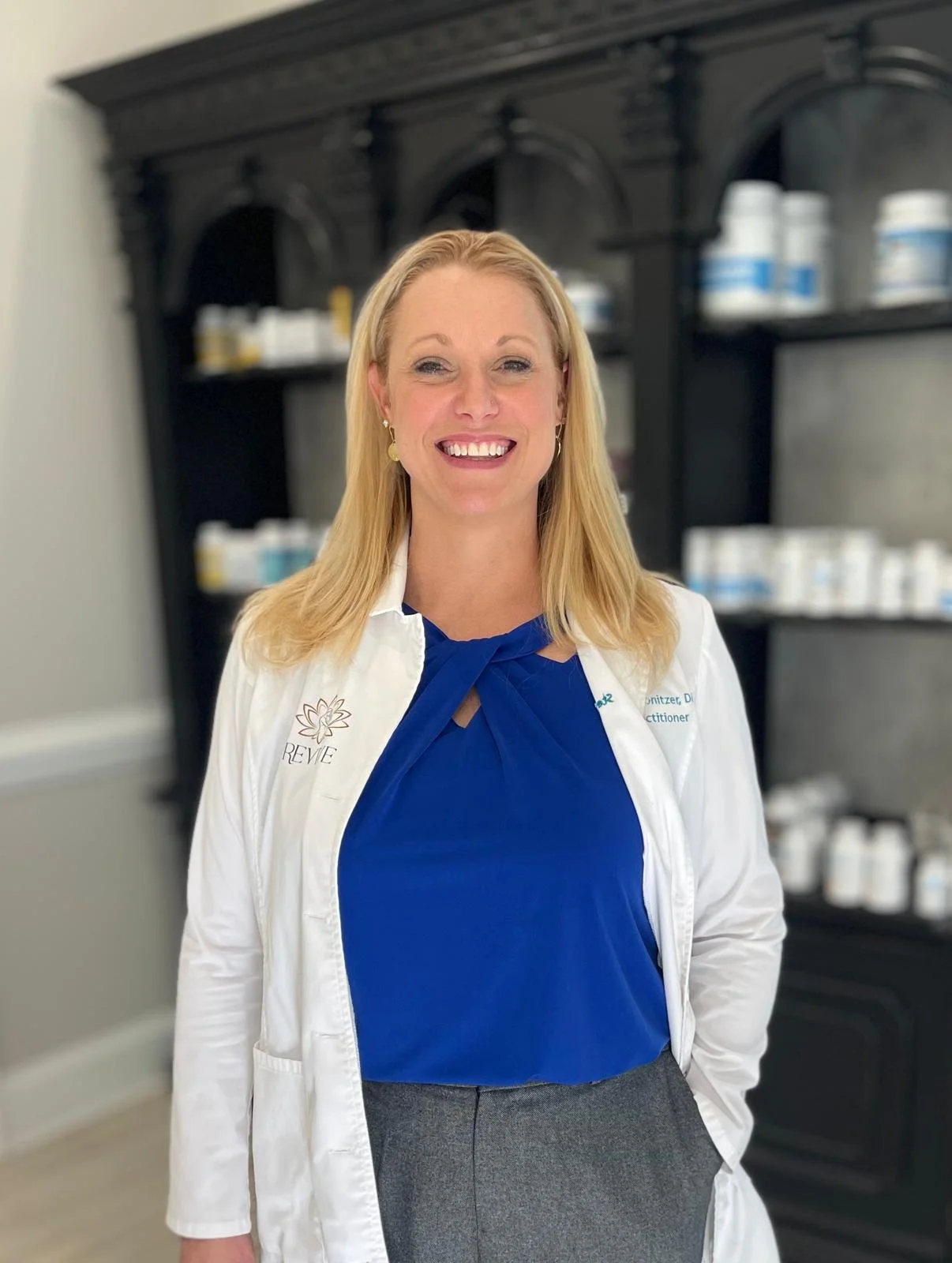 A smiling woman in a white lab coat and blue blouse standing in front of shelves of skincare products.