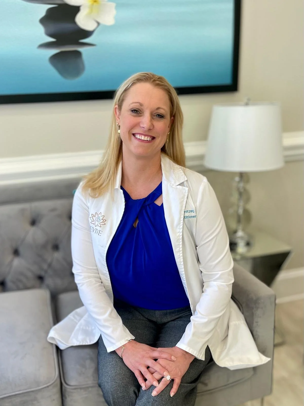 A smiling woman with blonde hair wearing a white coat and blue blouse sitting on a gray tufted sofa in a well-lit room with a lamp and a large framed artwork behind her.