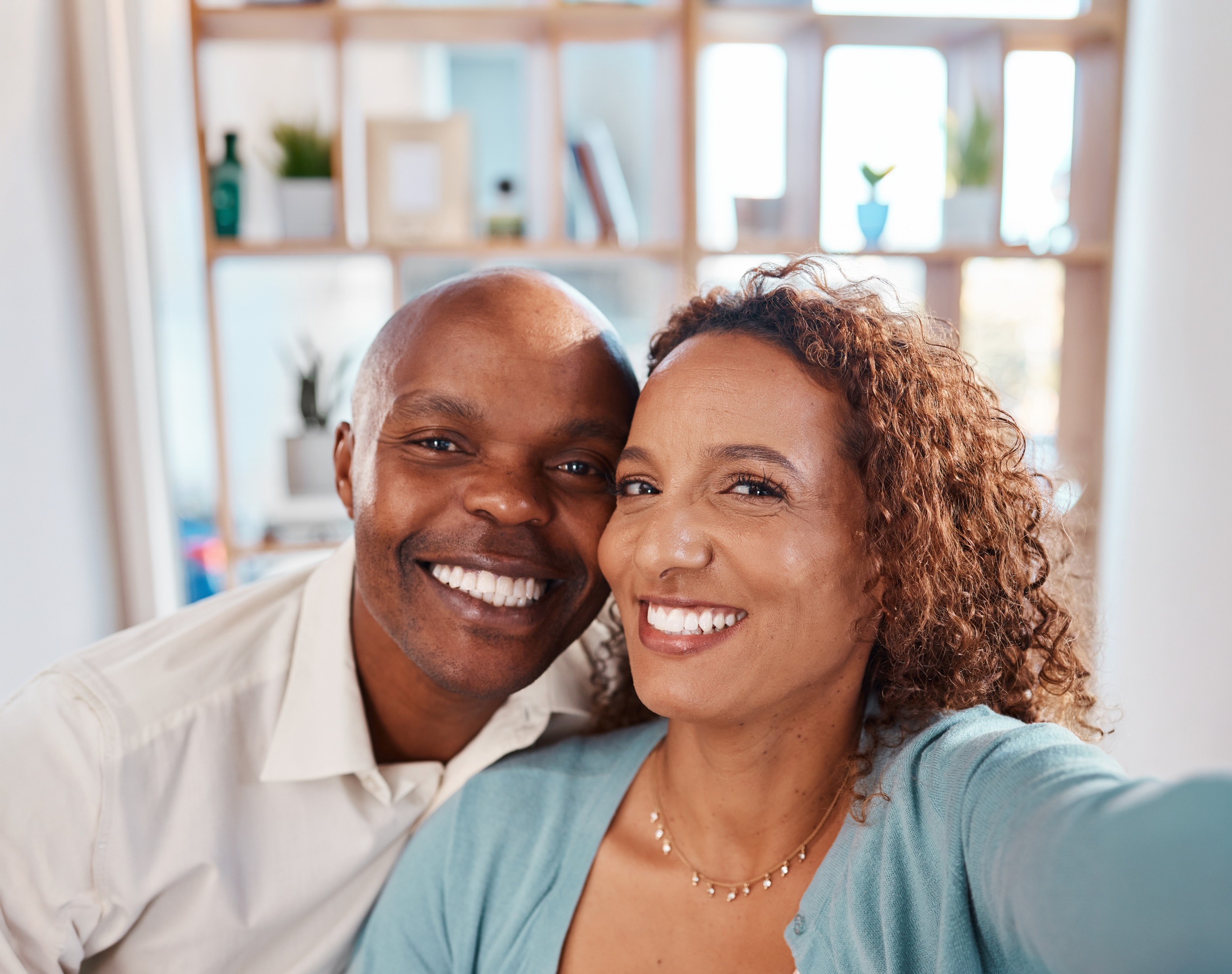 A smiling middle-aged Black couple taking a selfie in a well-lit room with a wooden shelf in the background.