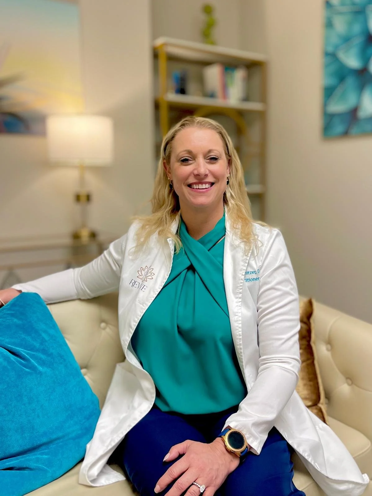 A woman in medical scrubs and a white coat sitting on a beige couch, smiling at the camera.