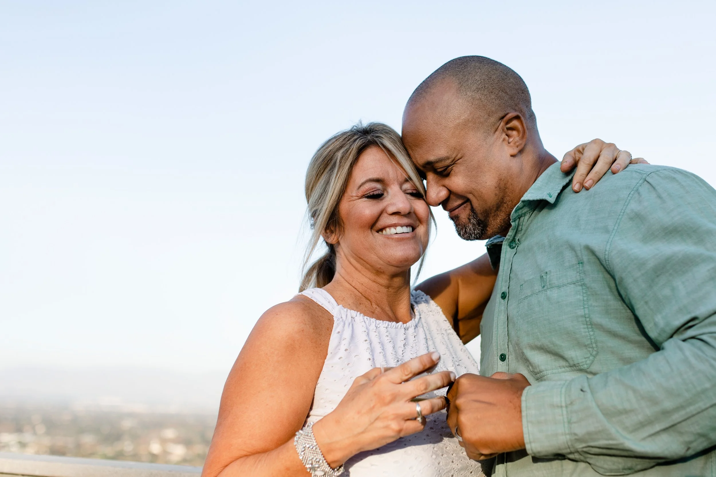 A smiling middle-aged woman and a young man are embracing on a rooftop with a cityscape in the background, touching foreheads with eyes closed.