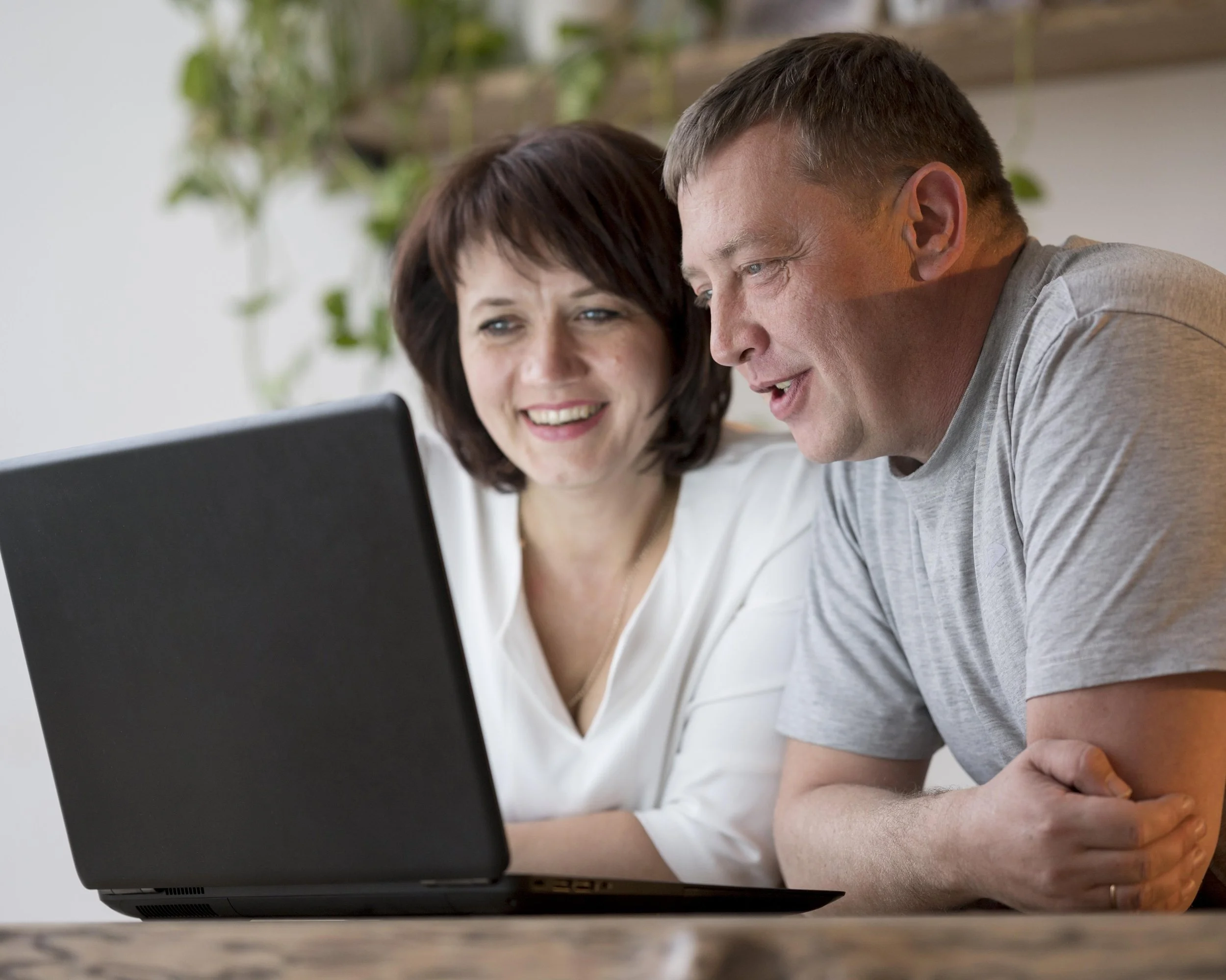 A man and woman sitting at a table looking at a laptop screen with smiles.
