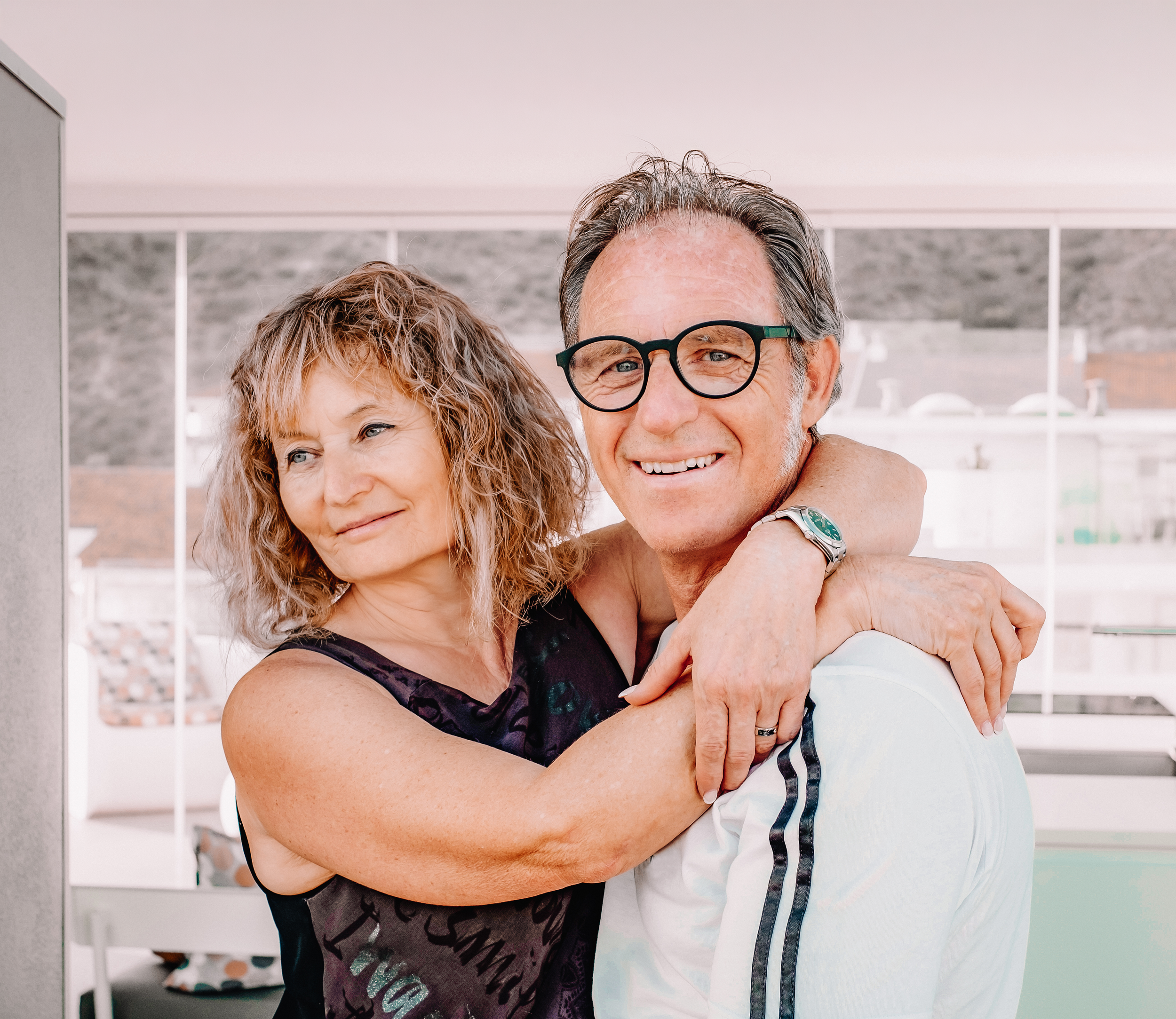 A happy middle-aged couple embracing each other in a bright room with large windows, with the man smiling and wearing glasses and the woman with curly hair looking at him affectionately.