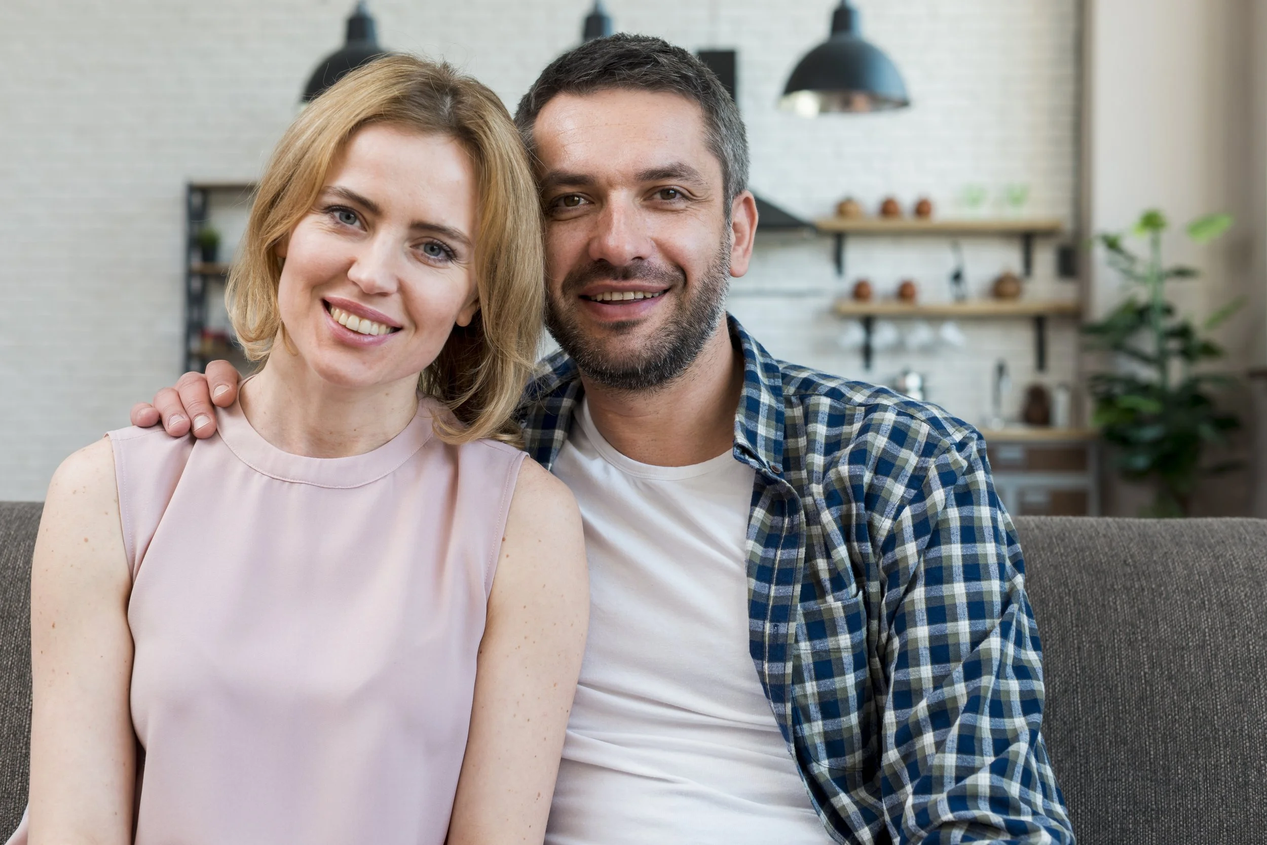 A smiling couple sitting on a couch in a cozy modern kitchen with white brick walls, black pendant lights, and wooden shelves with plants and decor.
