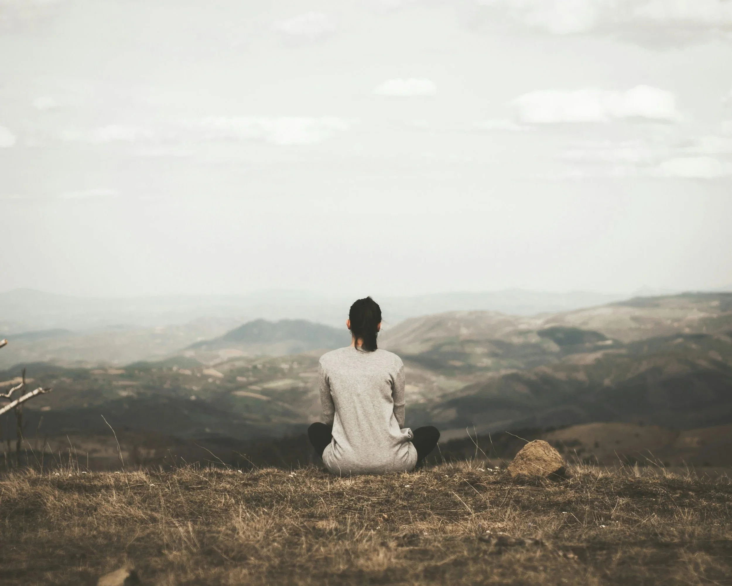 Person sitting cross-legged outdoors facing a vast landscape with mountains and cloudy sky.