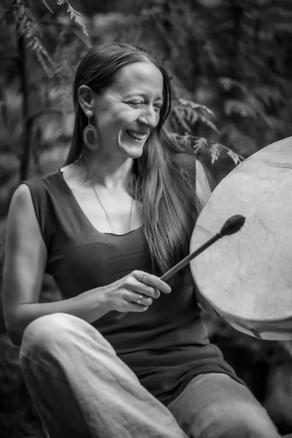 A woman joyfully playing a large hand drum outdoors surrounded by trees, wearing a sleeveless top, light-colored pants, and large earrings.