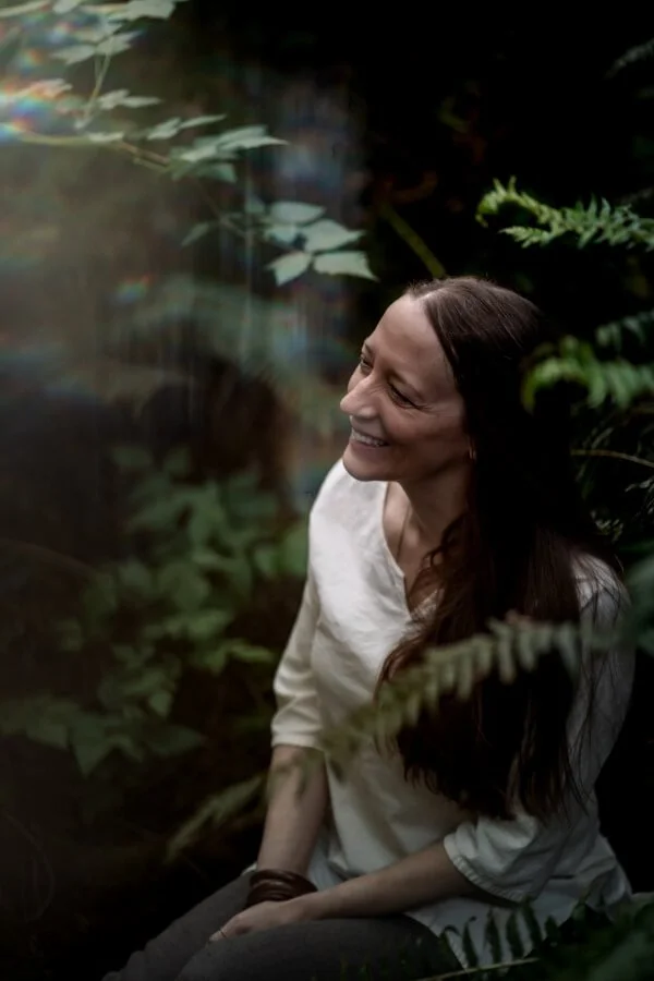 A woman with long dark hair and a white blouse sitting among green plants, smiling and looking to her right.