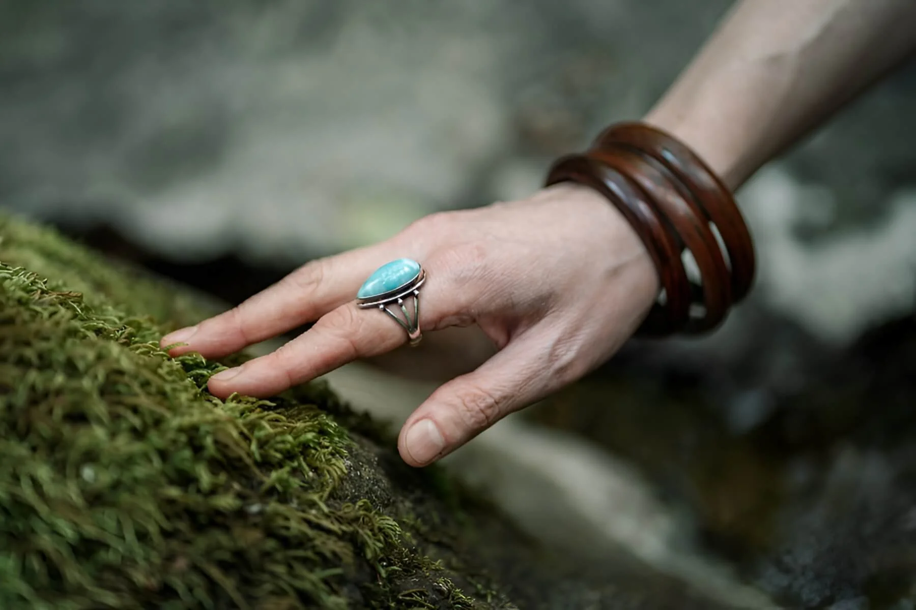A person's hand with a large turquoise ring and brown bracelets reaches toward moss-covered surface.