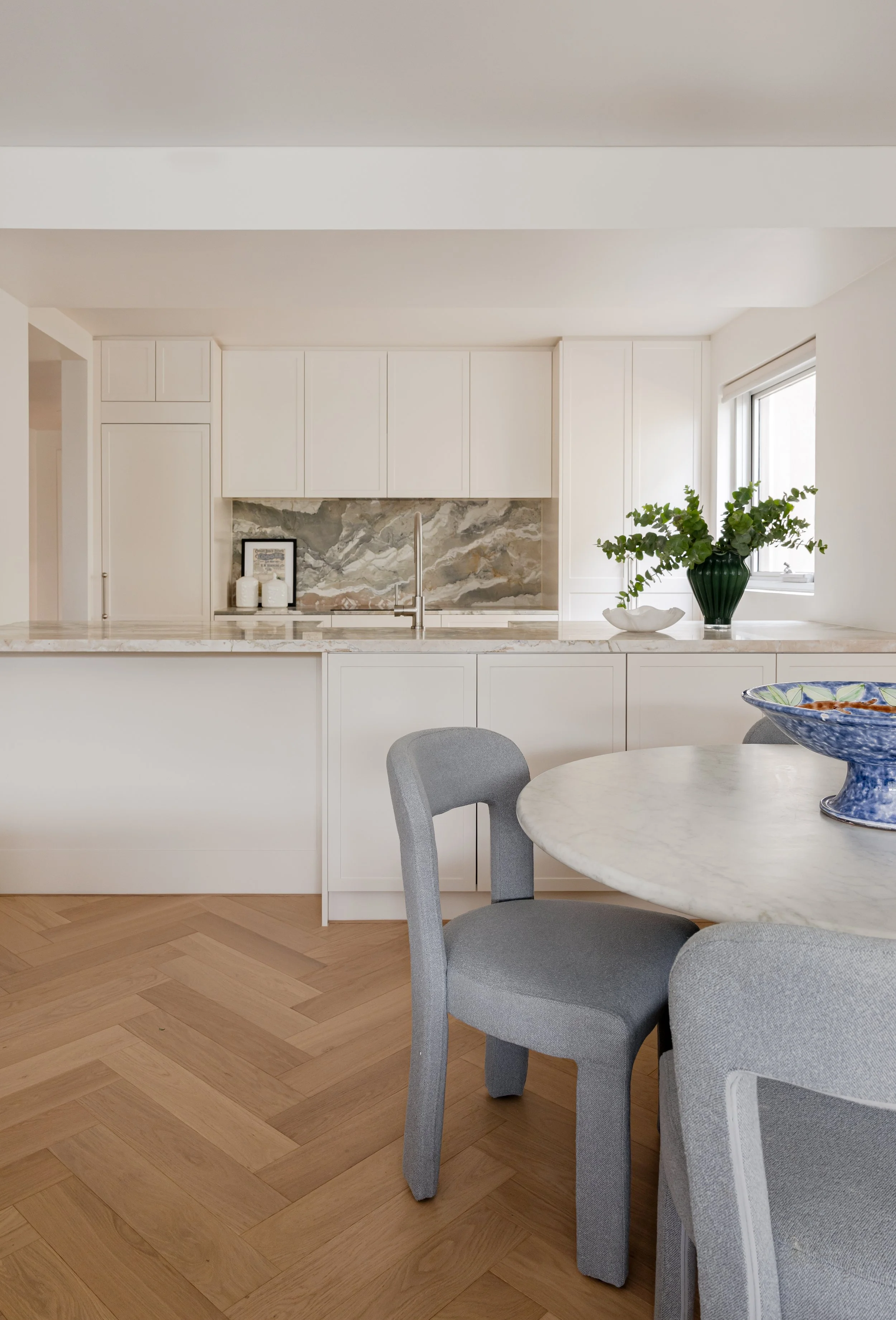 A modern kitchen with white cabinets, a marble backsplash, and a counter decorated with a framed picture, candles, a large green plant, and a white bowl. A round marble table with gray upholstered chairs is in the foreground, and the flooring is a light wood herringbone pattern.