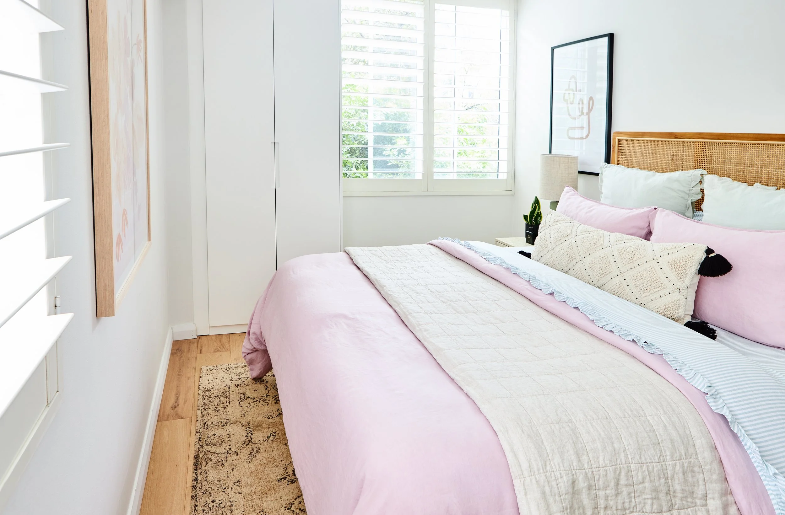 A cozy bedroom with a bed dressed in pastel pink and cream bedding, a rattan headboard, a nightstand with a lamp and a plant, a large window with white shutters, and framed art on the wall.