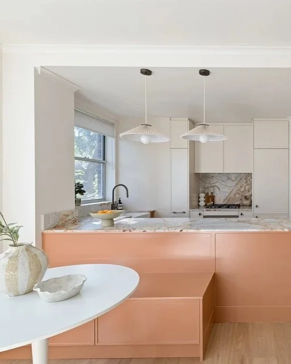 Modern kitchen with white cabinets, marble backsplash, pink storage unit, two hanging pendant lights, and a window with natural light.