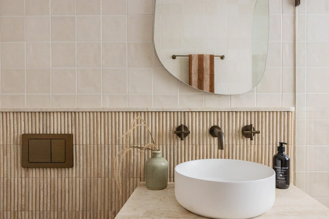 Bathroom sink area featuring a round mirror, beige tile wall, and a white vessel sink with a black soap bottle and a beige decorative vase on the countertop.