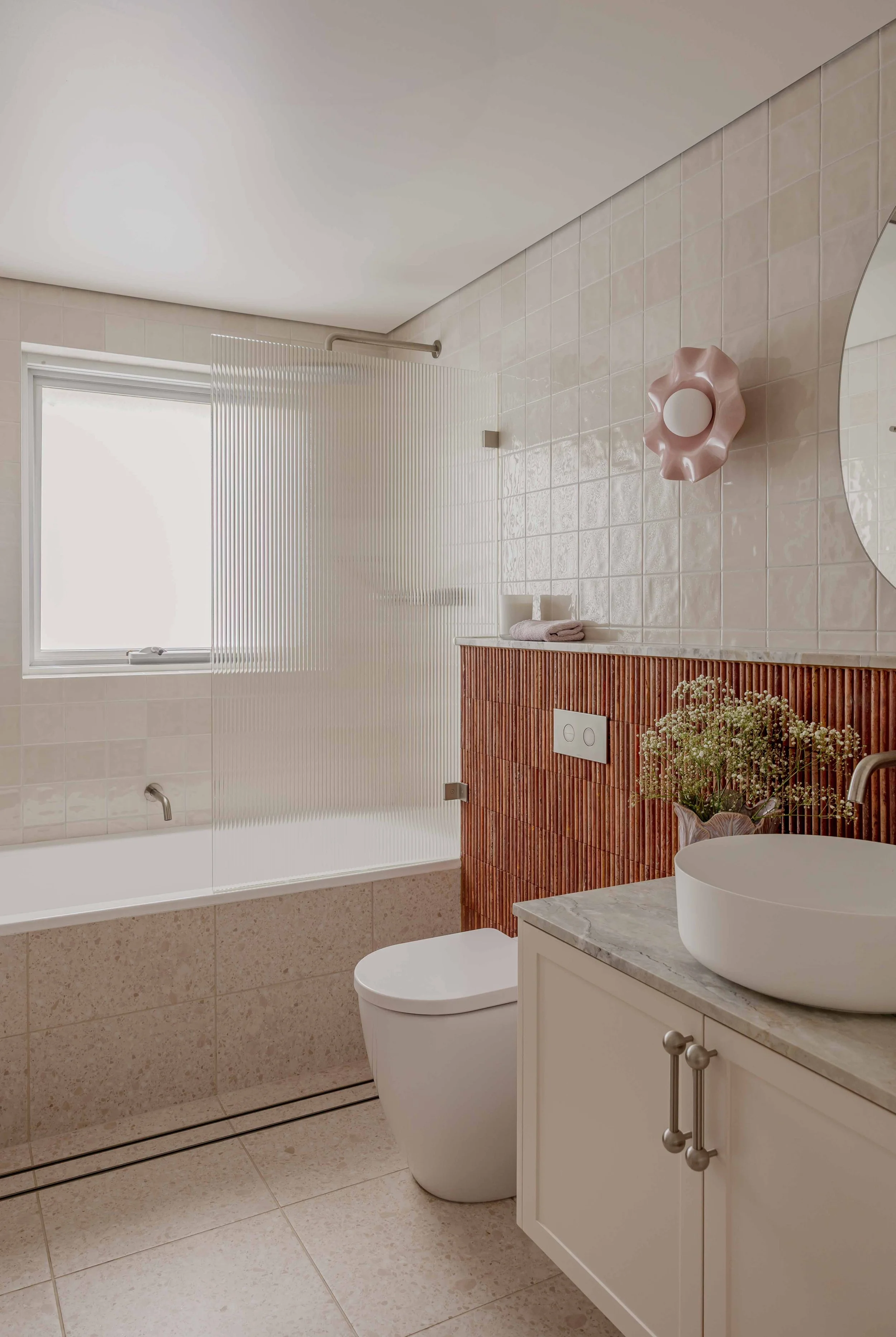 A modern bathroom with a frosted window, beige tiles, a white toilet, a tiled half-wall with vertical grooves, a marble countertop with a white vessel sink, a round mirror, a pink decorative wall piece, a vase with flowers, and a towel on the countertop.