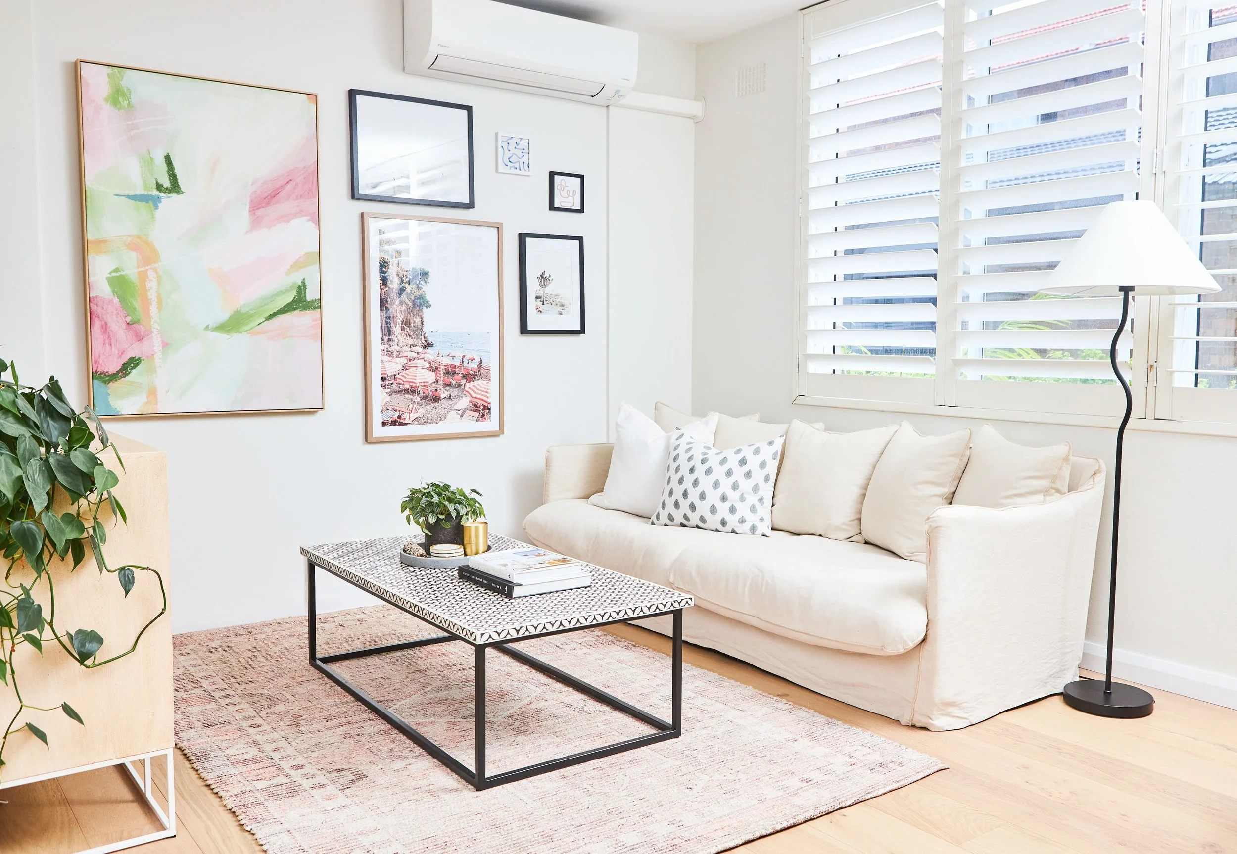 Bright living room with a white couch, a black and white patterned coffee table, a rug, wall art, a tall floor lamp, and a window with white blinds.