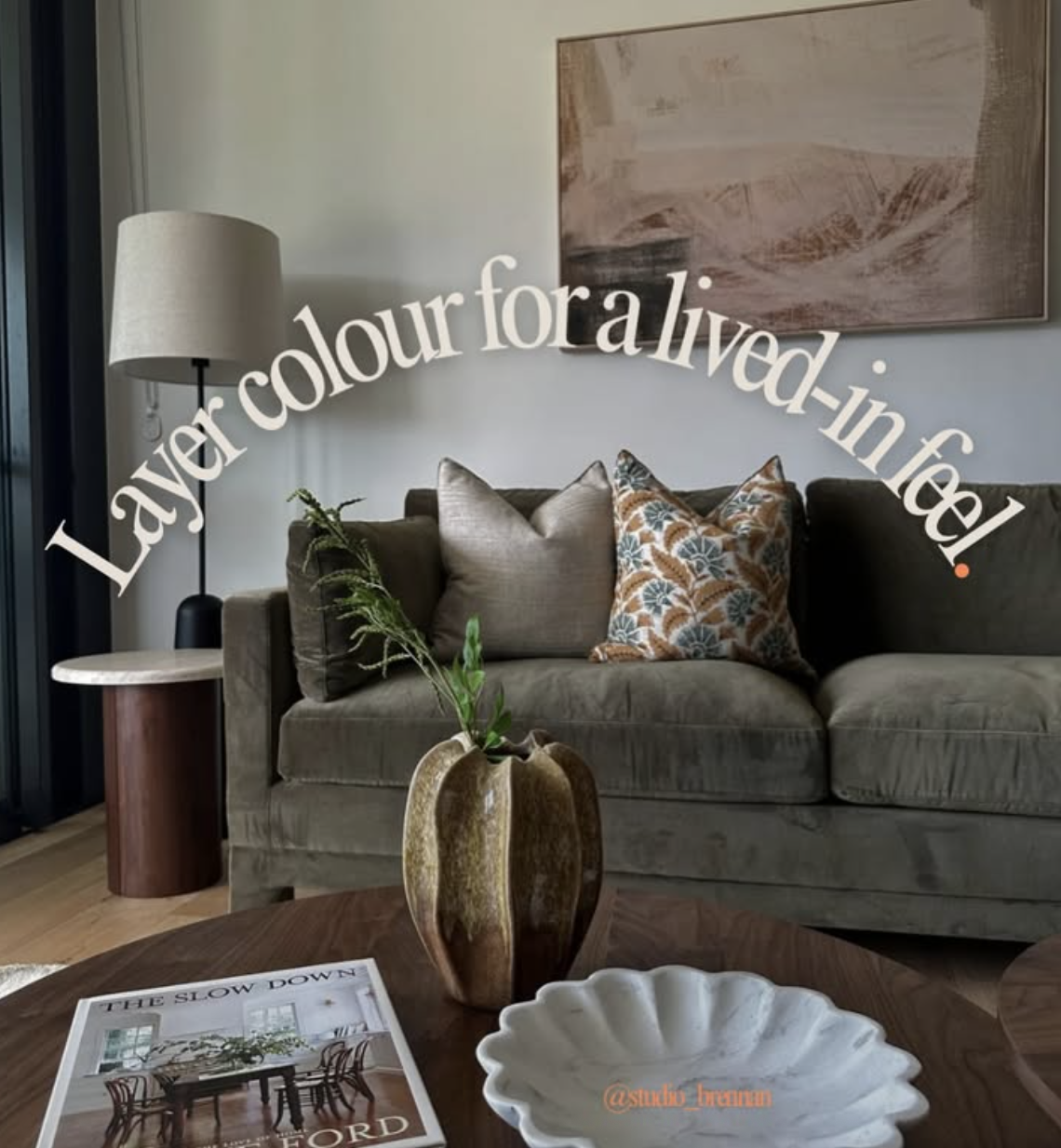 A living room with a gray sofa, decorative pillows, a wooden coffee table with a book and a ceramic dish, a tall vase with green plants, a white table lamp, and a large abstract painting on the wall.