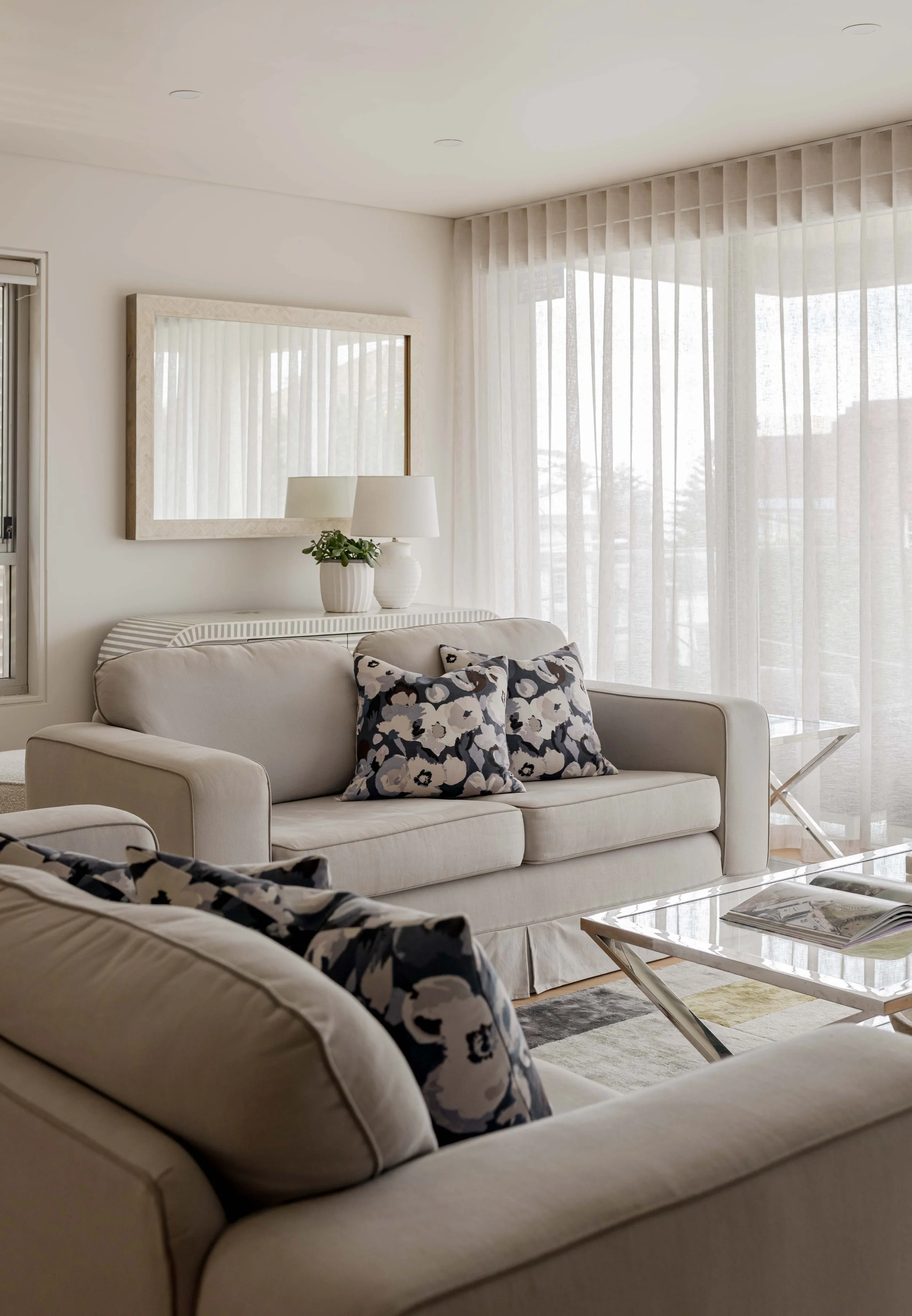A living room with a beige sofa with floral-patterned pillows, a glass-top coffee table with magazines, a white console table with ceramic vases and lamps, a large mirror on the wall, and sheer curtains allowing natural light.