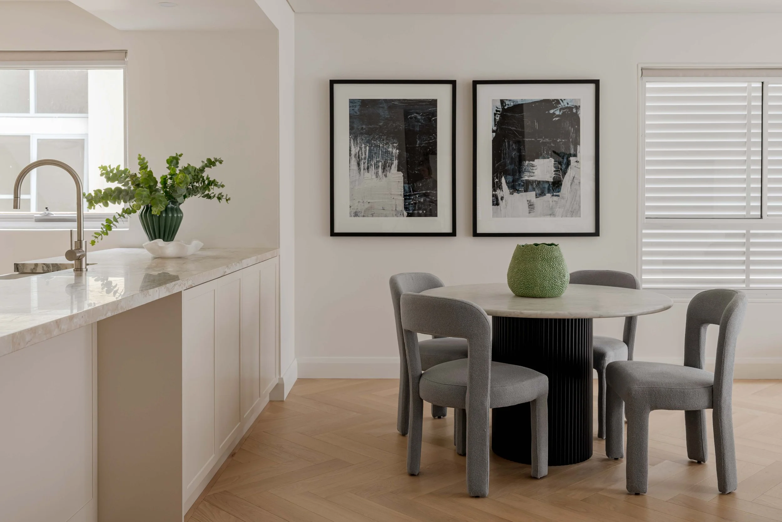 A modern kitchen and dining area with white walls, a marble countertop, a green plant in a green vase on the counter, black and white abstract artwork on the wall, a round marble dining table with four gray upholstered chairs, and a green textured vase on the table. There's a window with white blinds on the right.