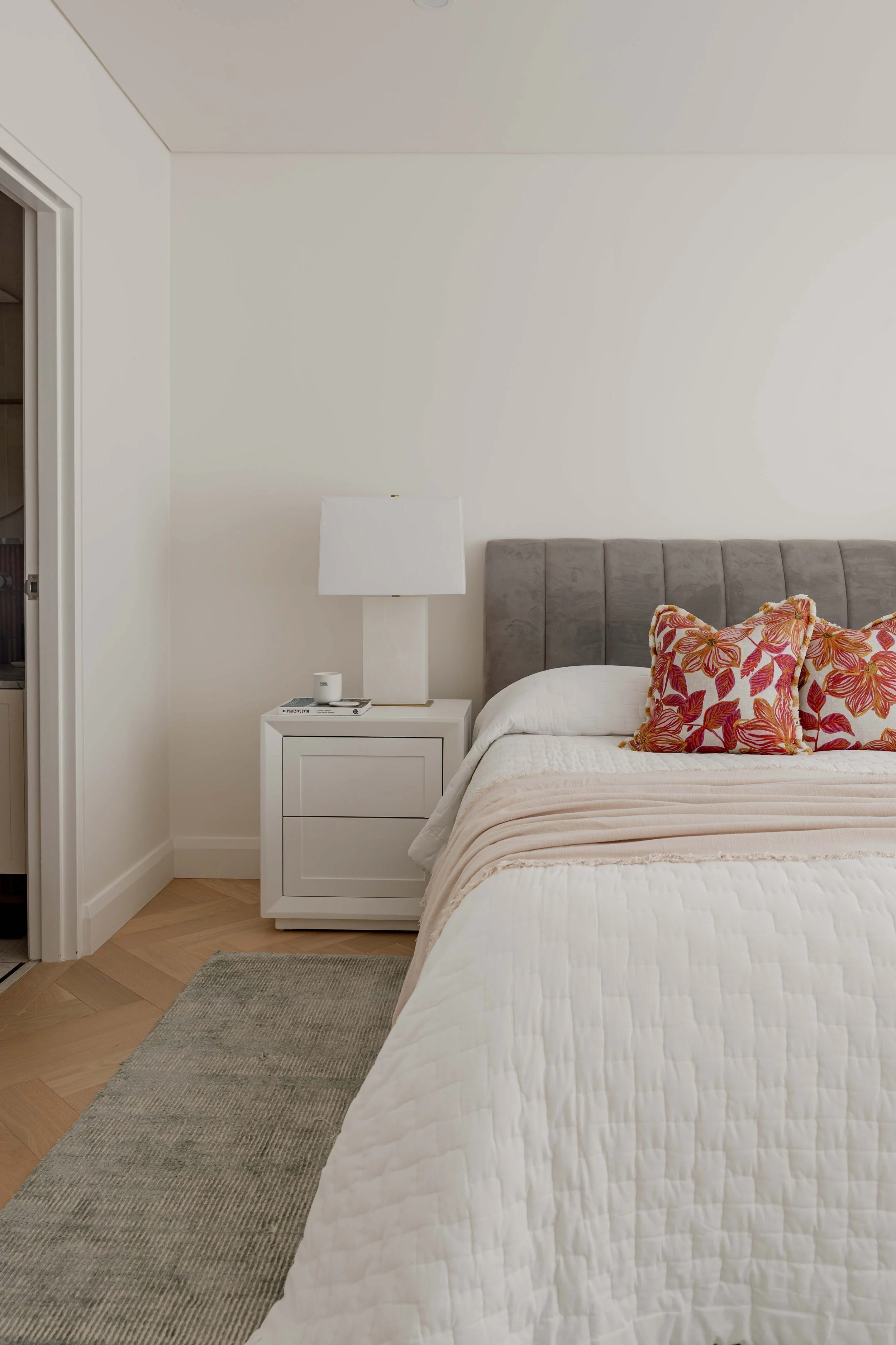 A bedroom with a bed, a gray upholstered headboard, a white nightstand with a lamp, a candle, and a magazine, two decorative pillows with a red and orange floral pattern, a beige quilted blanket, a gray rug, and a doorway leading to another room.