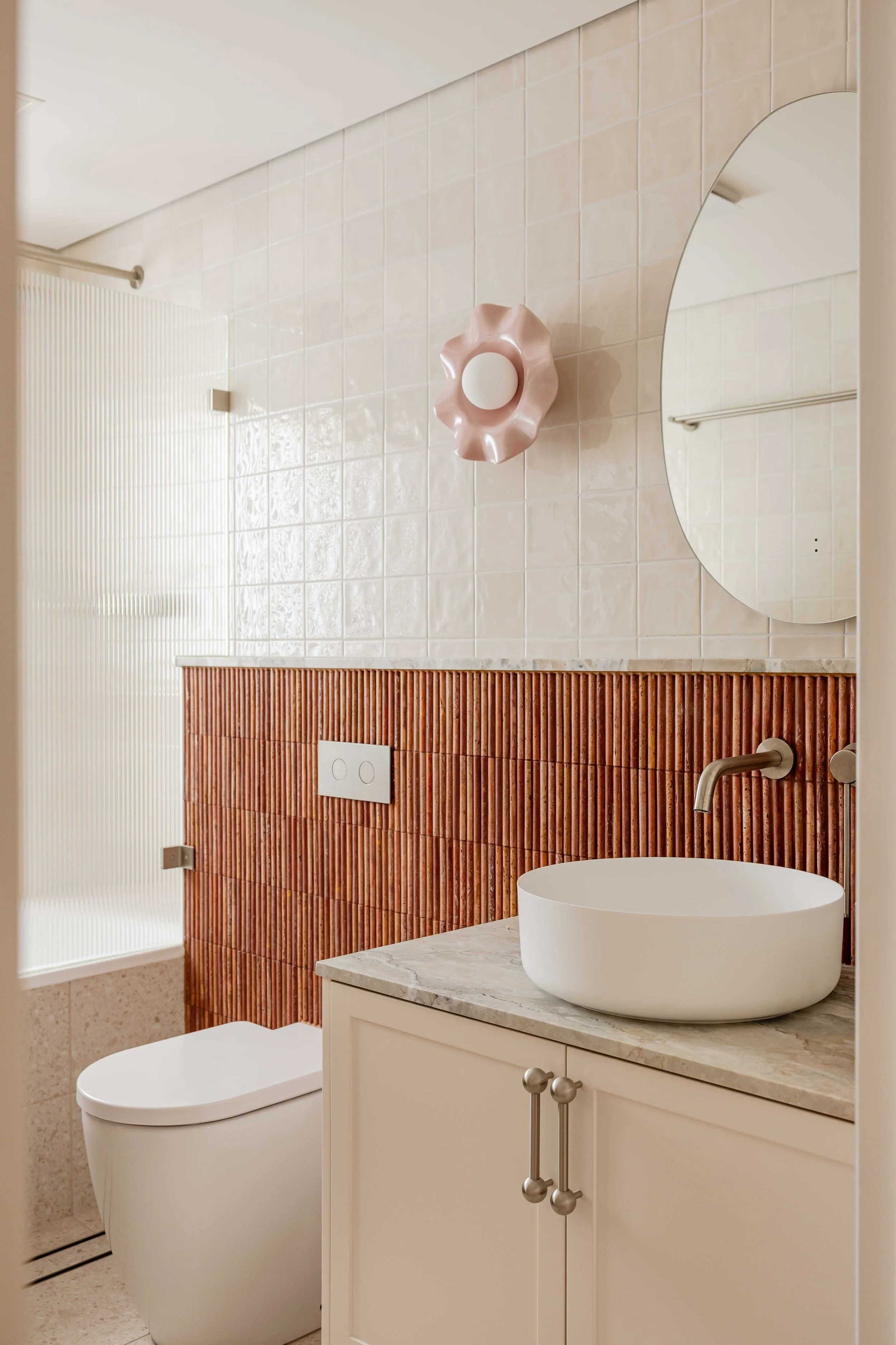 Modern bathroom with beige tiled walls, a white toilet, a white vessel sink on a beige cabinet with marble countertop, round mirror, pink decorative wall light, and textured brown tiled backsplash.