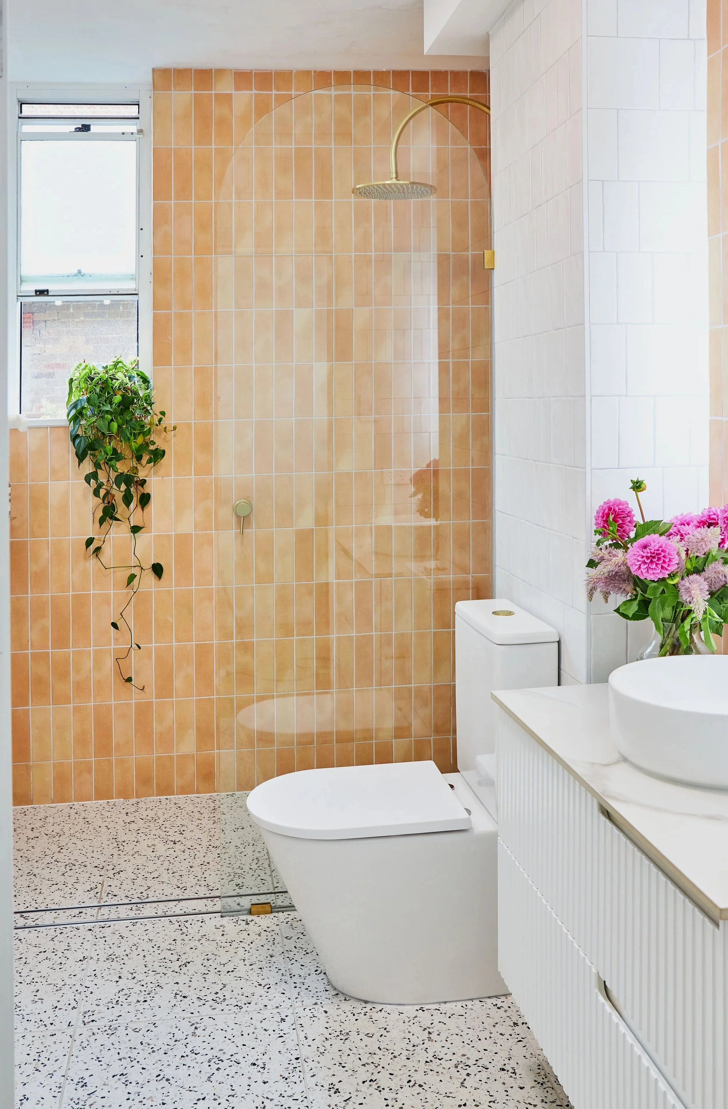 Modern bathroom featuring a walk-in shower with a gold showerhead and a glass partition, a white toilet, a white vanity with a vessel sink, and pink flowers in a vase. There is a window with a green plant beside it and speckled flooring.