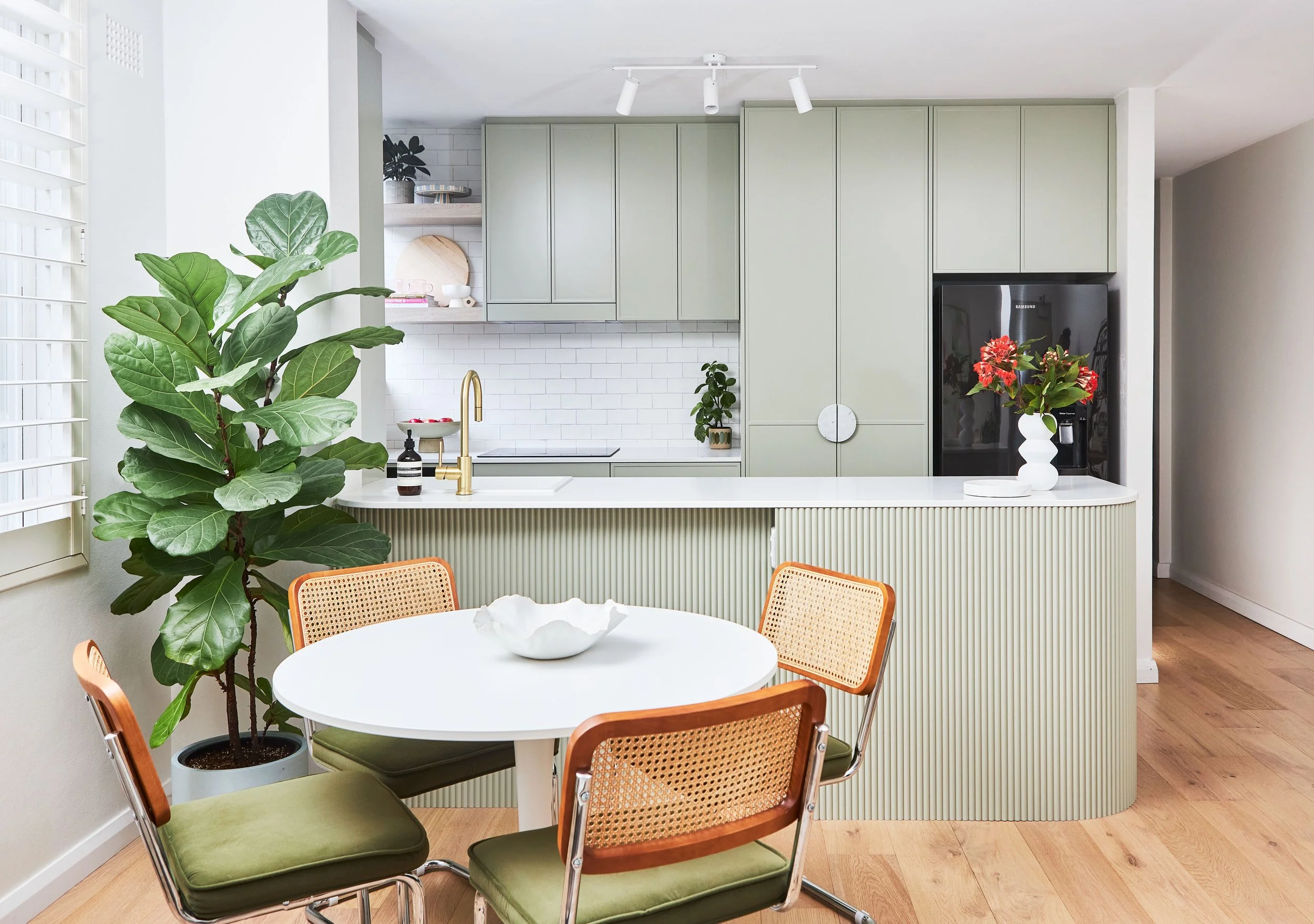 Modern kitchen with light green cabinets, white backsplash, and a white island with ribbed detail. A black refrigerator with a flower arrangement, a round white dining table with four chairs, and a large leafy plant near a window with blinds.