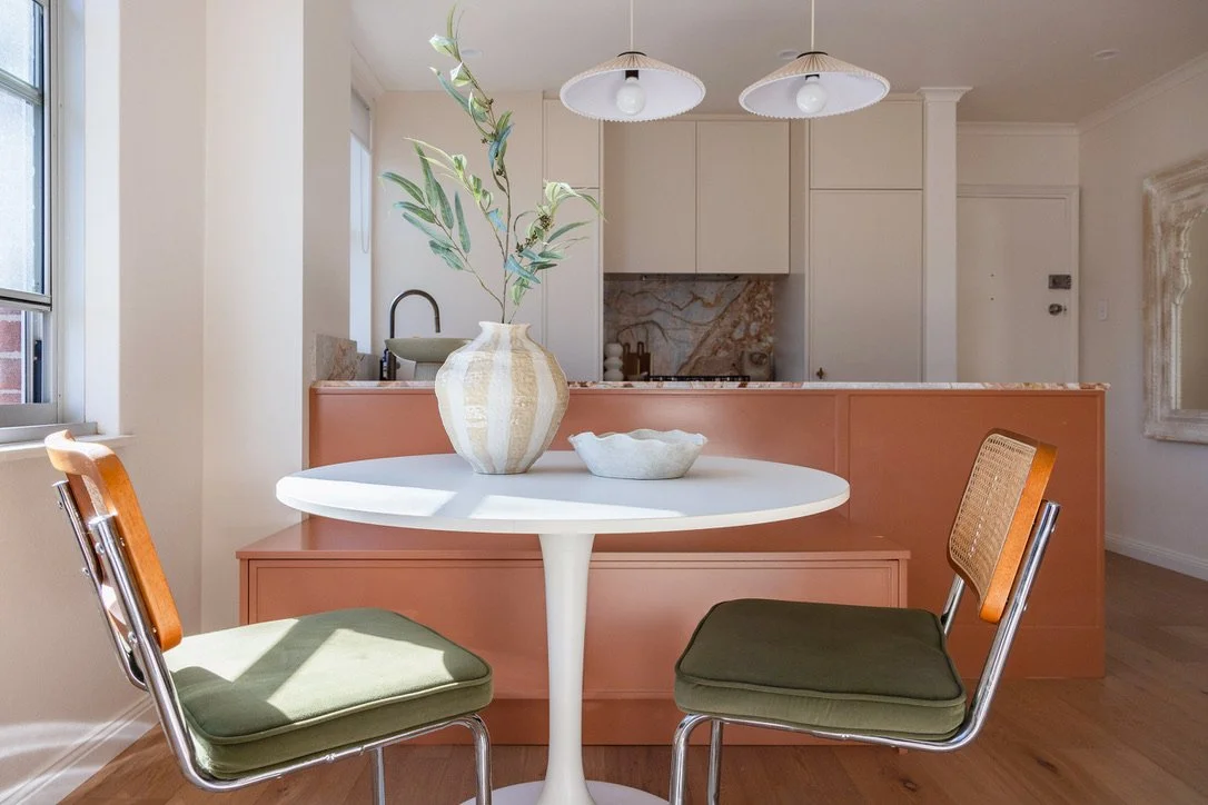 A round white dining table with two green-cushioned chairs in a bright room, overlooking a kitchen with beige cabinets, a marble backsplash, and a pinkish-brown partition. A large vase with green foliage is on the table.