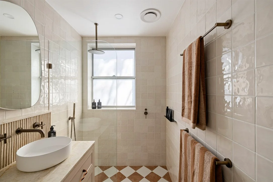 Modern bathroom with beige tiled walls, a window, a walk-in shower with a rain showerhead, a white vessel sink, and brown towel racks with towels.