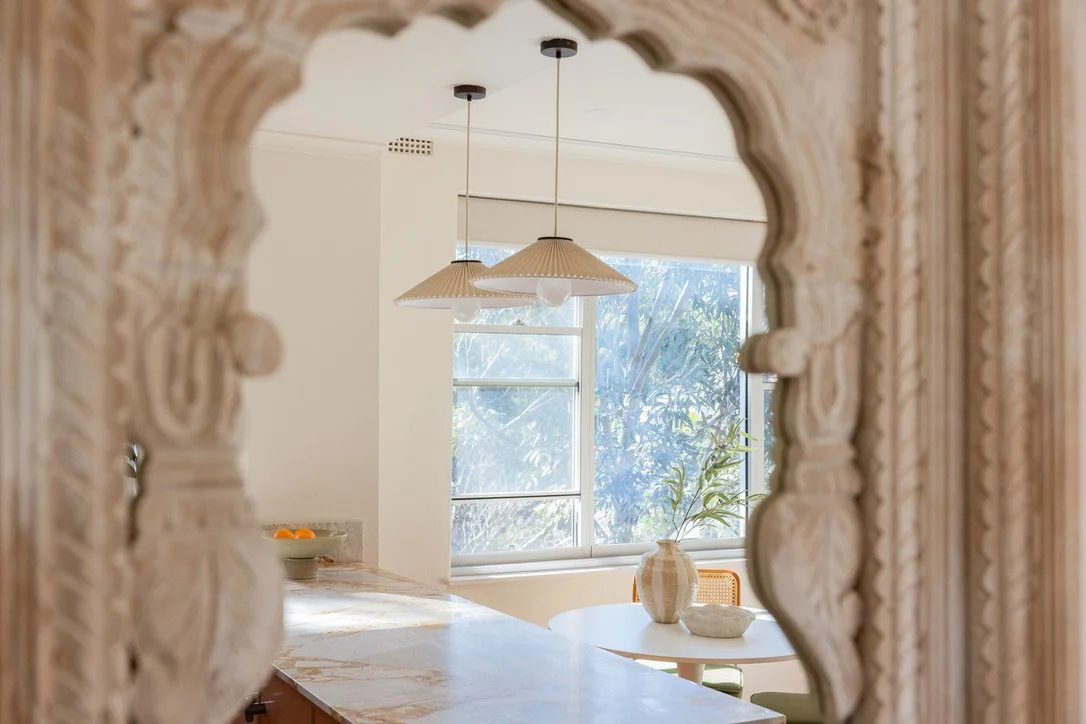 View through an ornate mirror showing a modern kitchen with a marble countertop, a round dining table, pendant lights, and a large window with greenery outside.