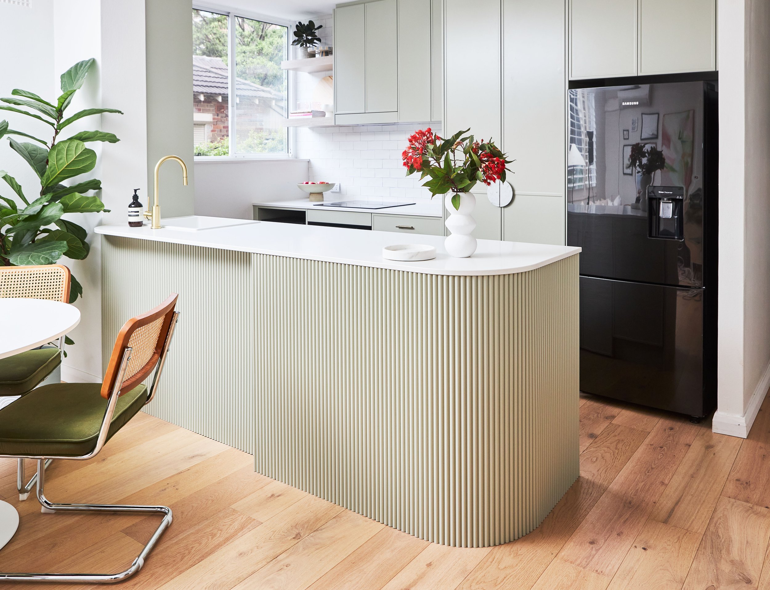 Modern kitchen with white cabinetry, a black refrigerator, and a white island with a gold faucet. There are plants and red flowers in a white vase on the island, with a window letting in natural light and a view outside.