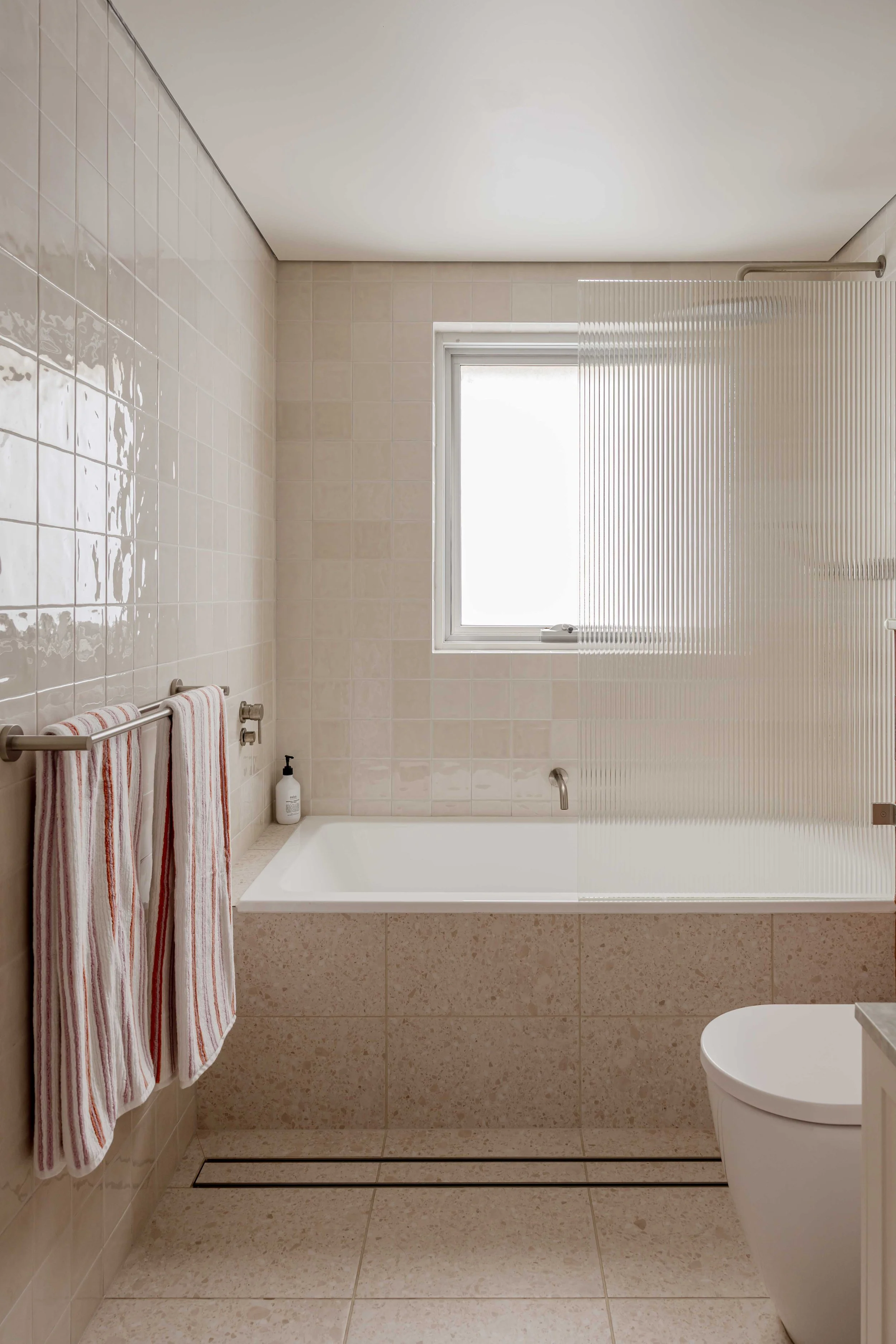 A minimalist bathroom with beige tiled walls and floor, a bathtub with a partial privacy screen, a window, and two wall-mounted towels.