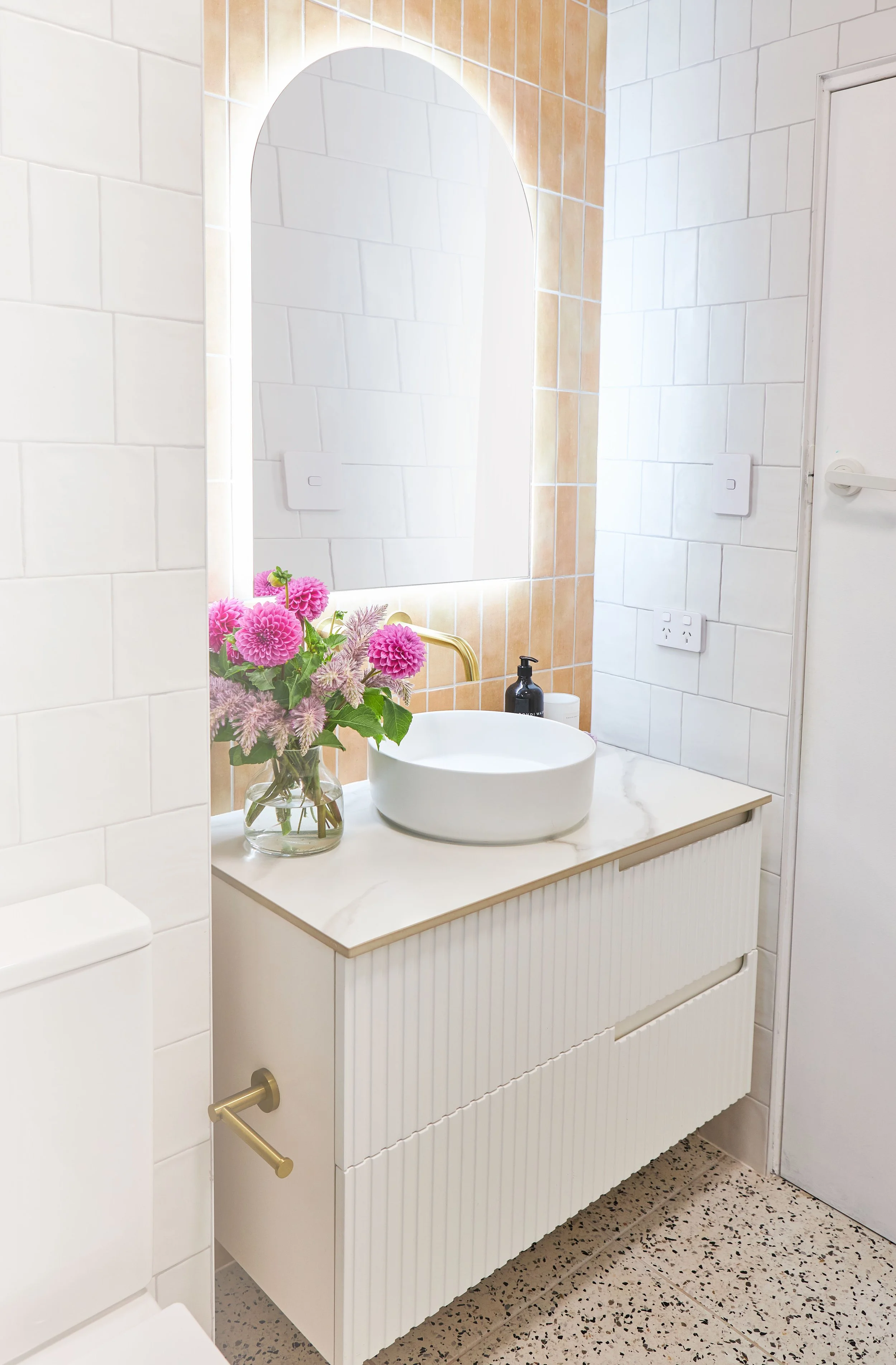 Bathroom with a white vanity, marble countertop, round vessel sink, gold faucet, large mirror with backlighting, pink flowers in a vase, soap dispenser, and a closed white door.