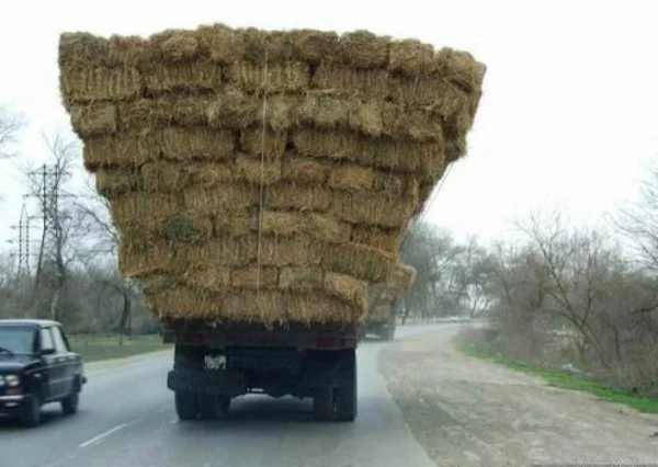 A truck transporting a large load of hay bales on a rural road, with a car passing by.