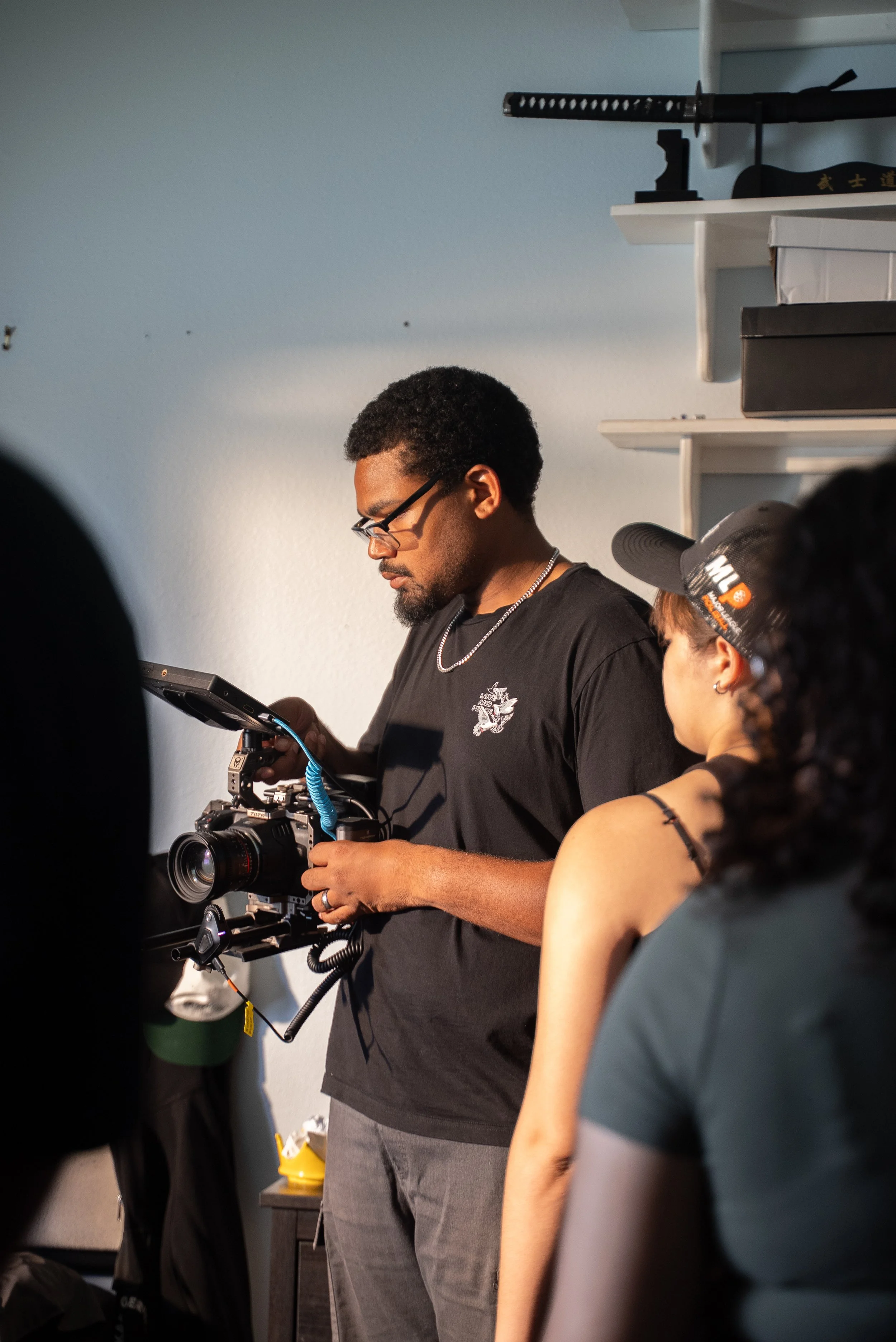 Man operating a professional camera, with a woman and part of another person's head in the foreground, in a room with shelves and a sword on display.