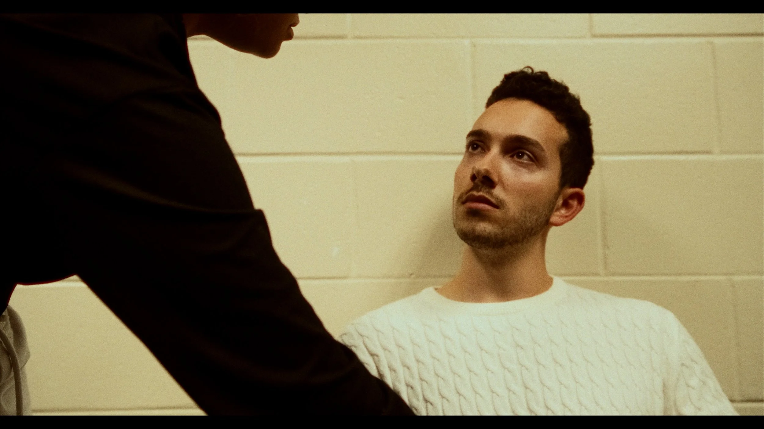 A young man with dark hair and light skin sitting against a beige cinderblock wall, looking up with a serious or concerned expression while another person, partially visible and wearing a dark jacket, leans over him.