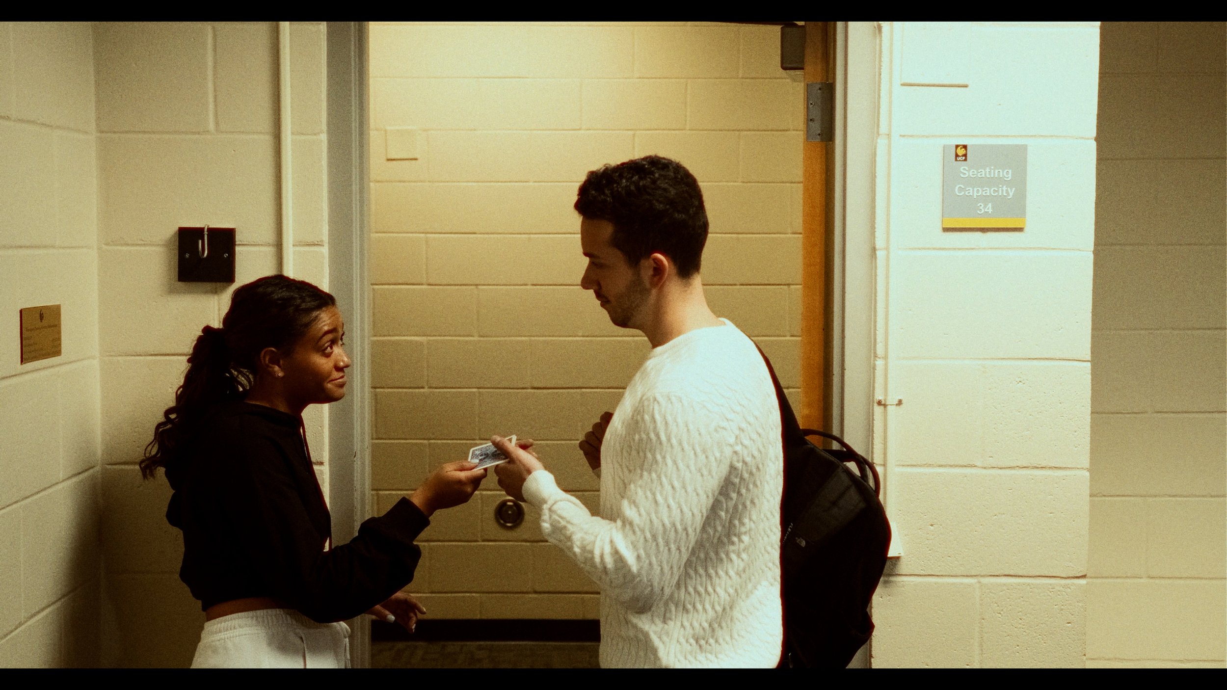 A young woman and young man are standing in a hallway near an elevator. The woman, with curly hair in a ponytail, is handing the man a card. The man has dark hair, is wearing a white sweater, and carries a backpack.