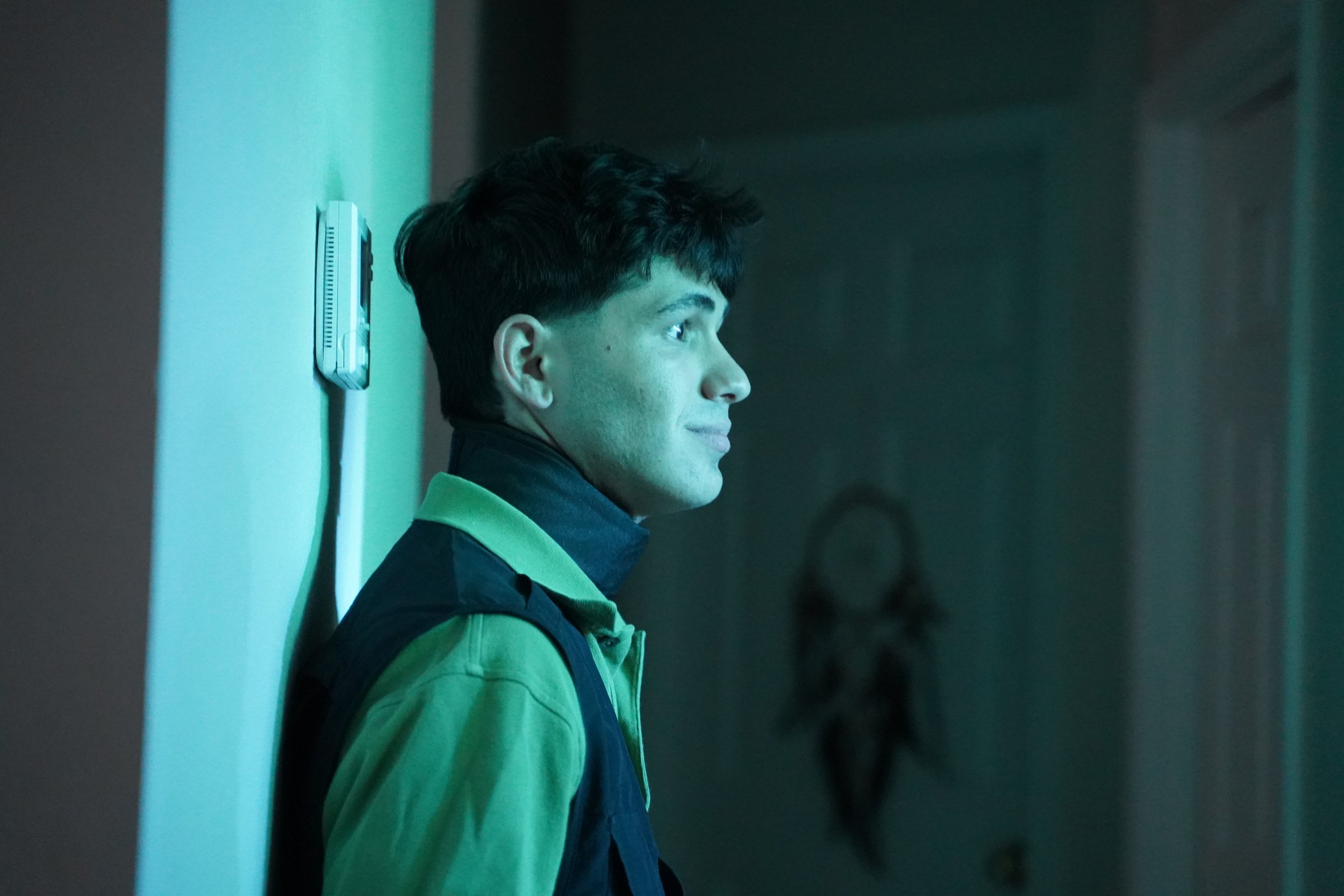 A young man with dark hair, wearing a green shirt with a black vest, stands in profile with his head leaned against a wall and a slight smile, illuminated by teal-colored lighting.