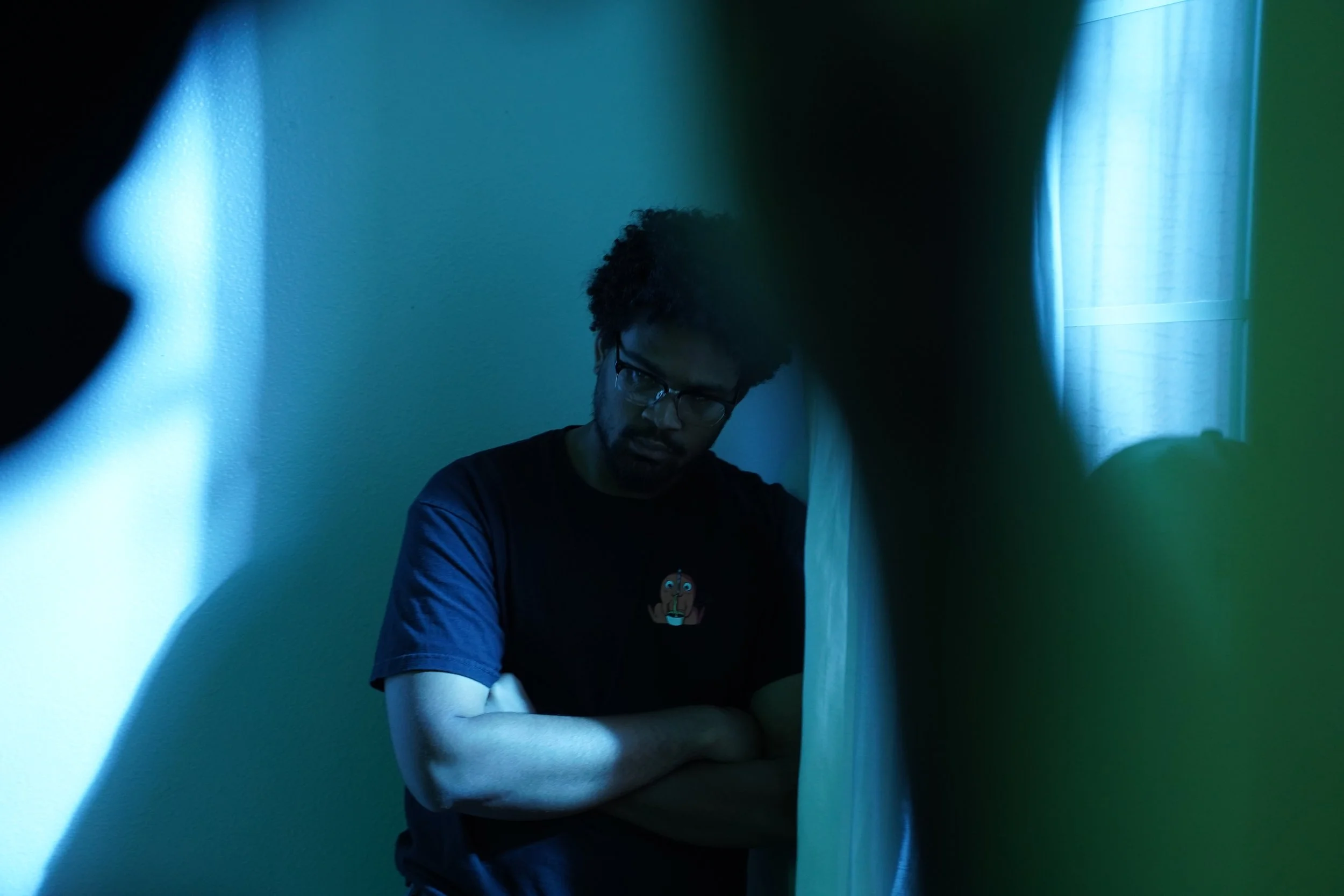 A young man with glasses and curly hair standing in a dimly lit room, with blue and green light reflections on the walls, seen through an irregular frame or object in the foreground.
