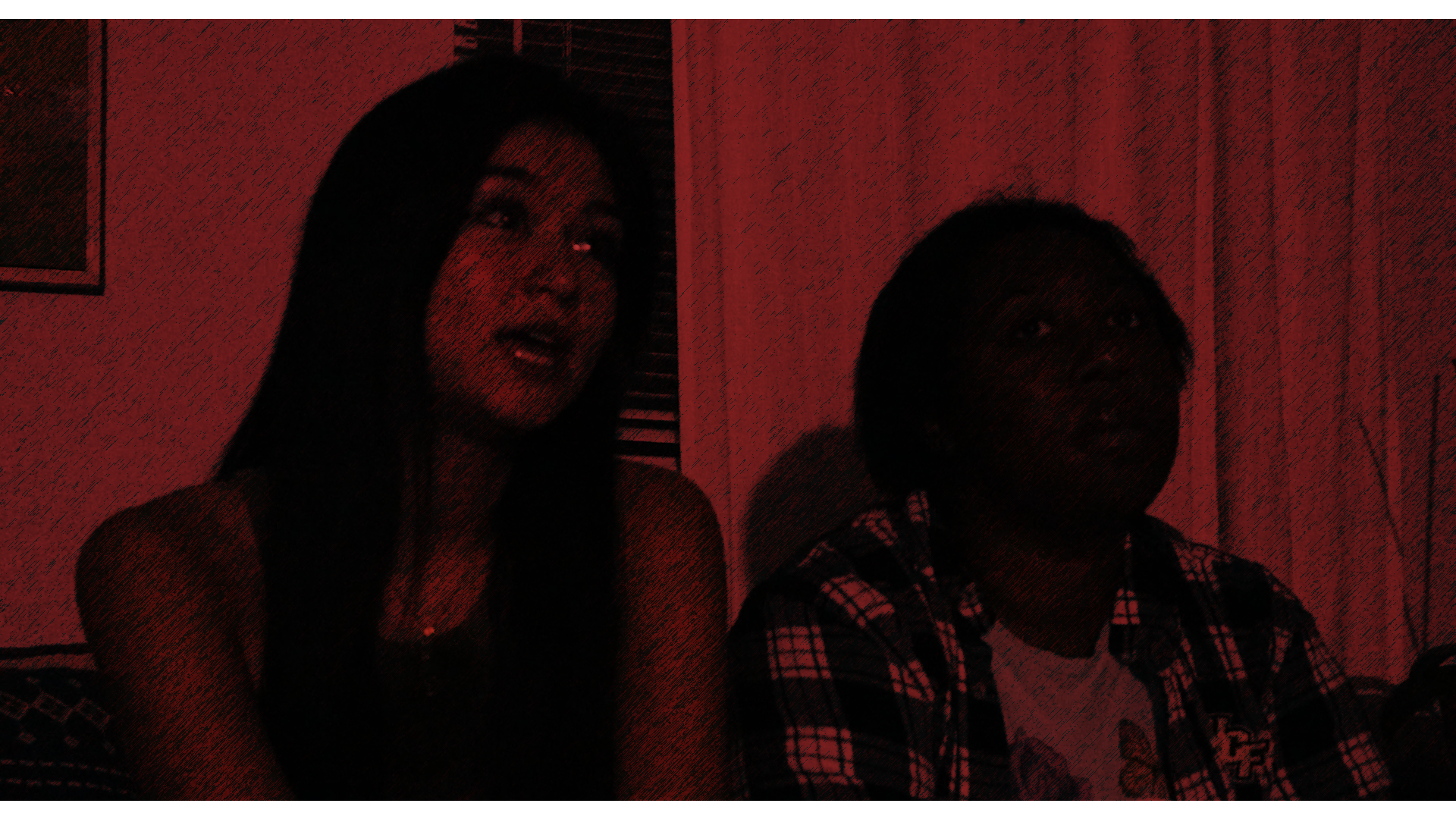 A woman with long black hair and a man with short black hair sitting indoors in a room with pink curtains and a framed picture on the wall. The lighting is dim and red-tinted.